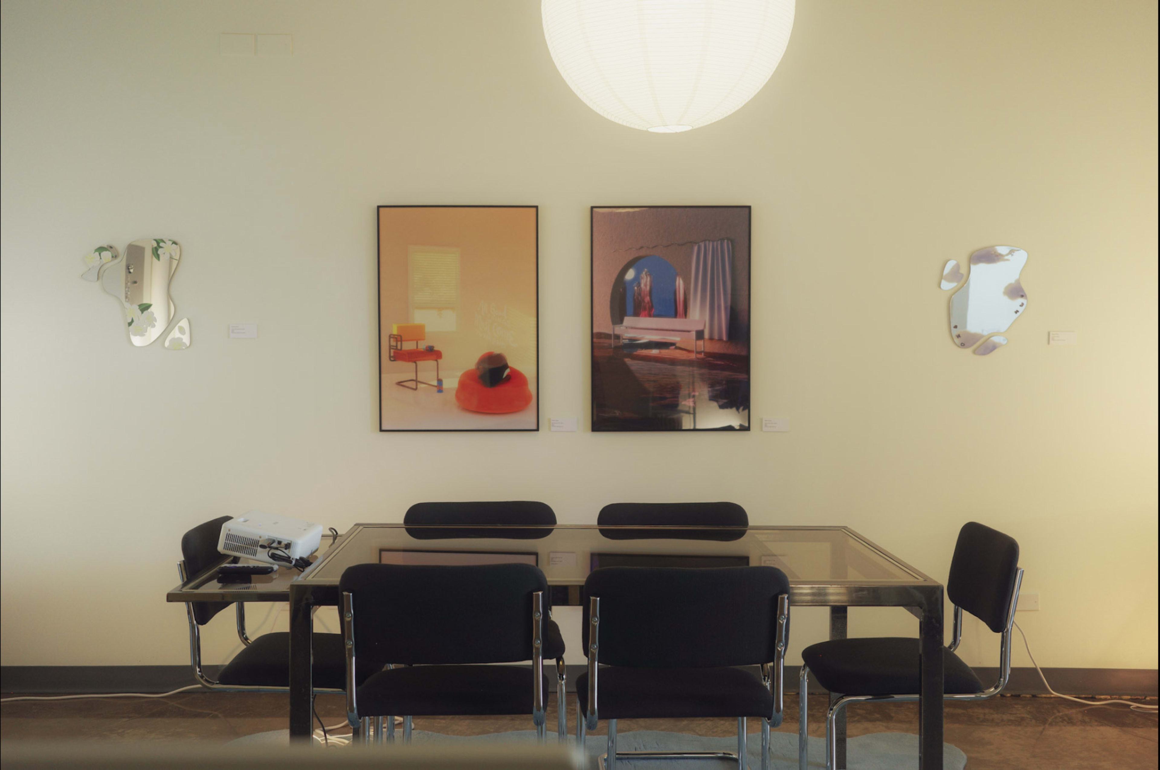 The image shows a modern dining area featuring a glass table surrounded by black chairs, with two framed artworks and a large hanging light on the wall.