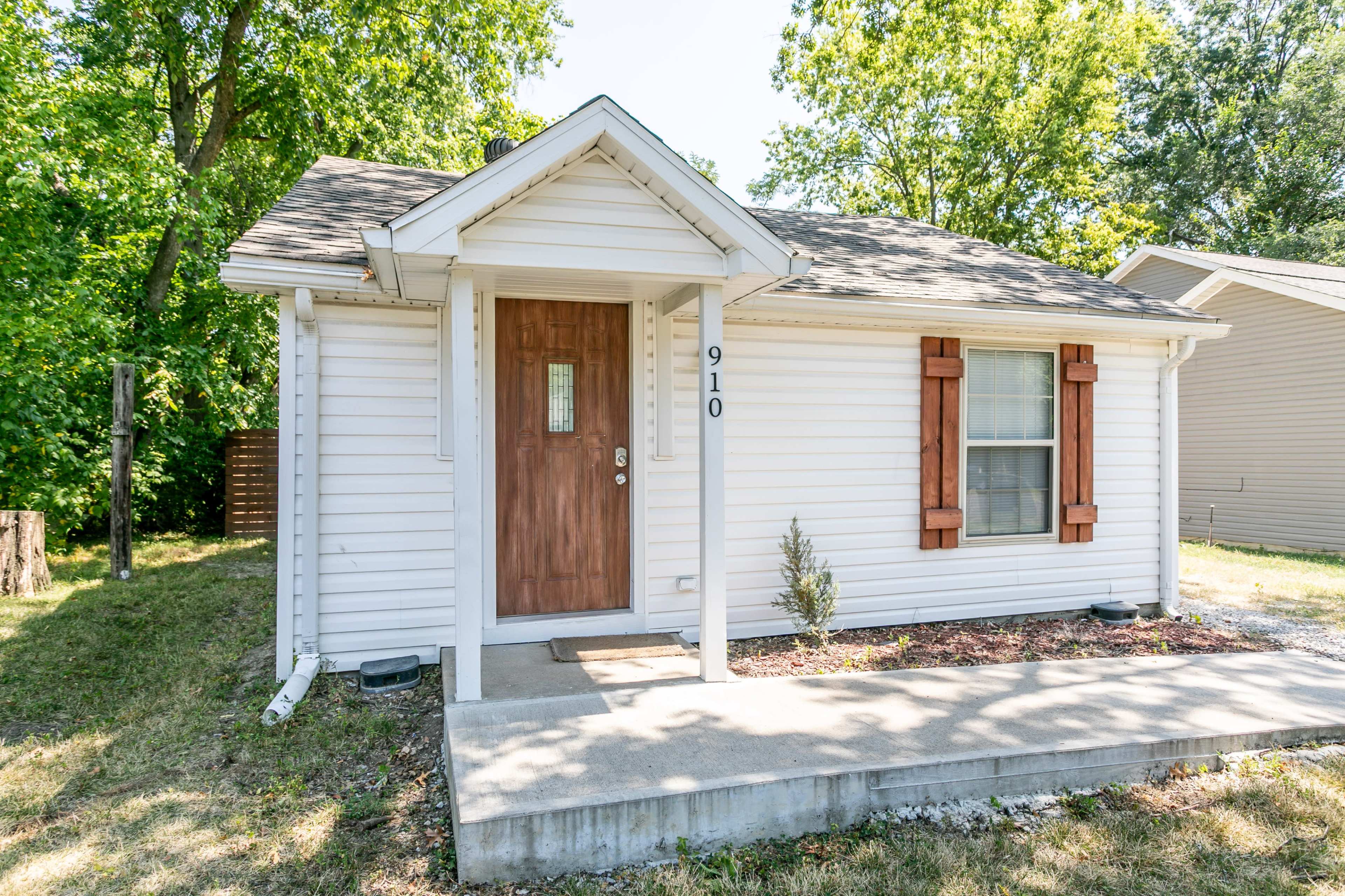 A small, single-story house with a front porch, wooden door, and shutters on the windows, surrounded by green trees and grass.