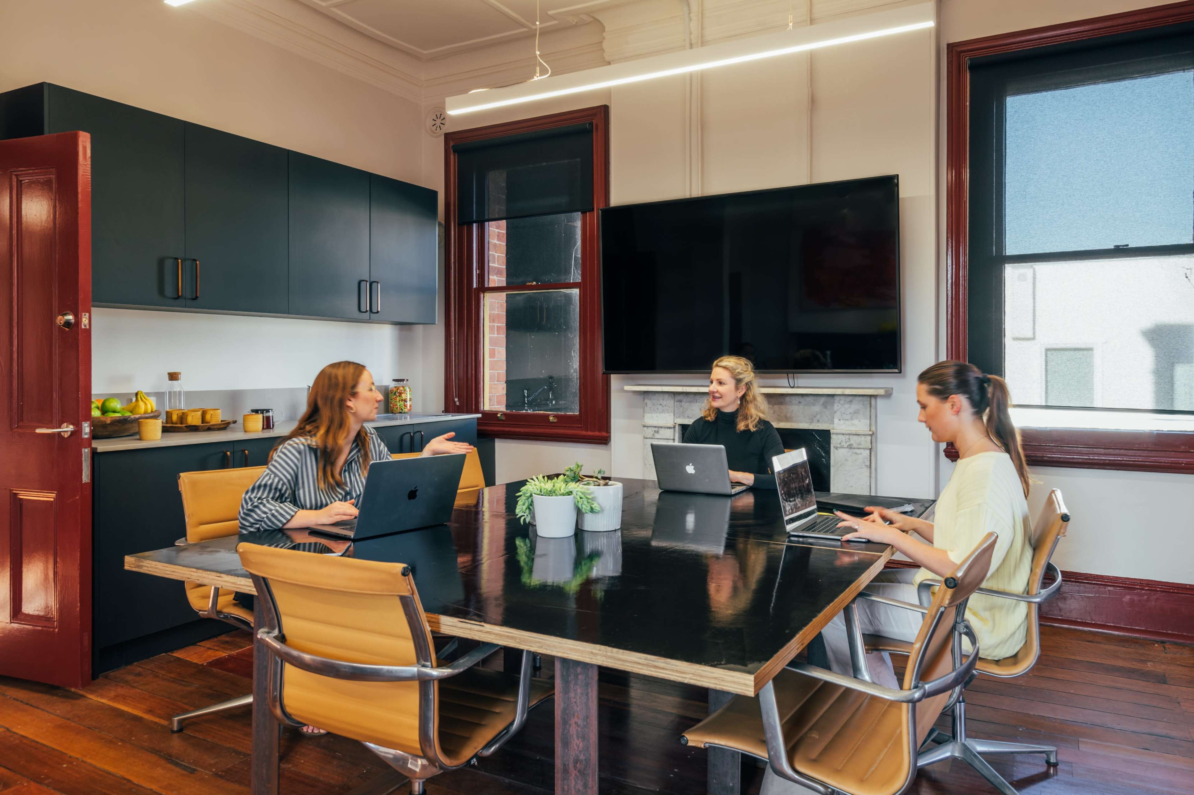 Three women are seated at a conference table in a modern office with laptops, discussing while a large screen displays a view of the room.