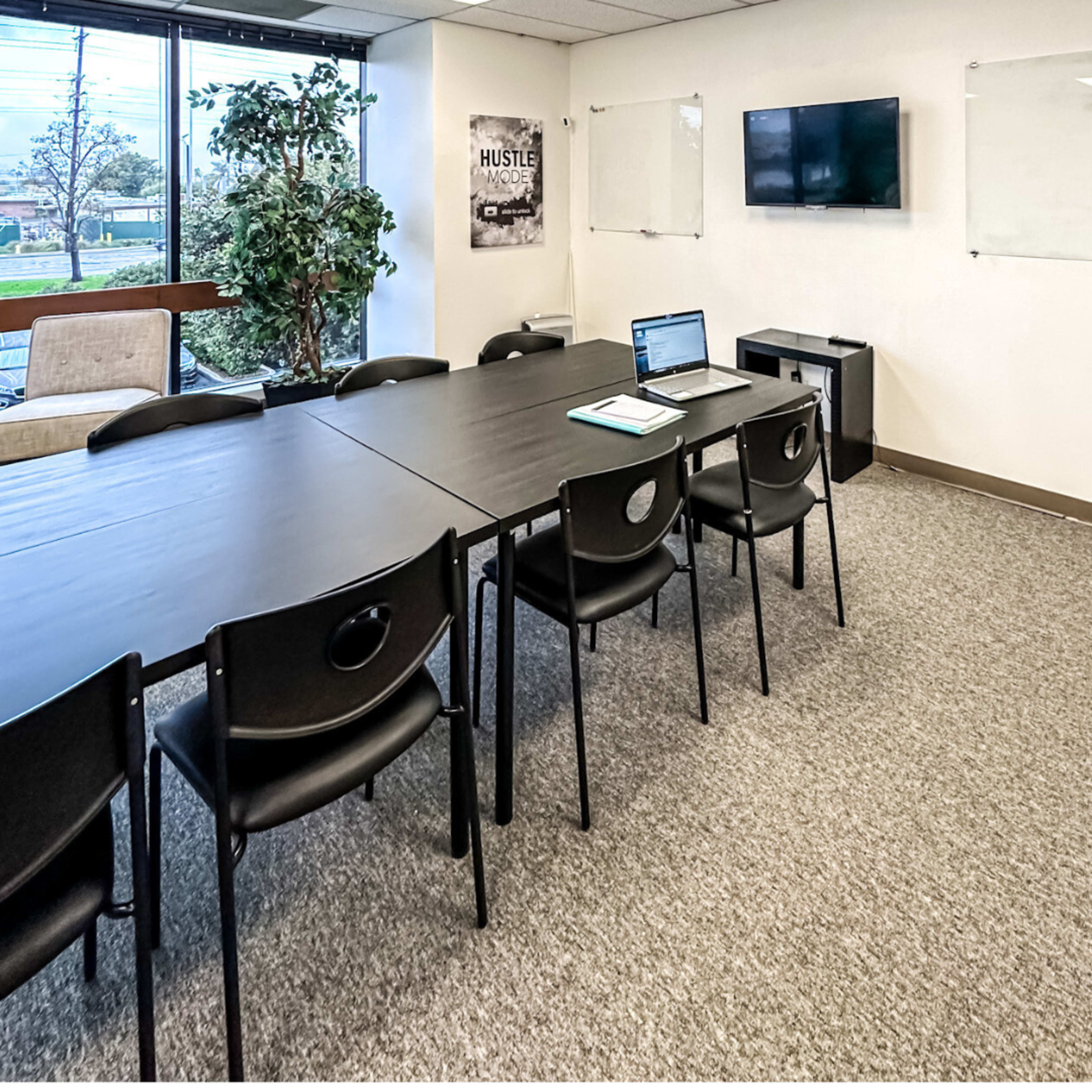 A modern conference room features a long black table surrounded by chairs, with a laptop open on the table and a television mounted on the wall.