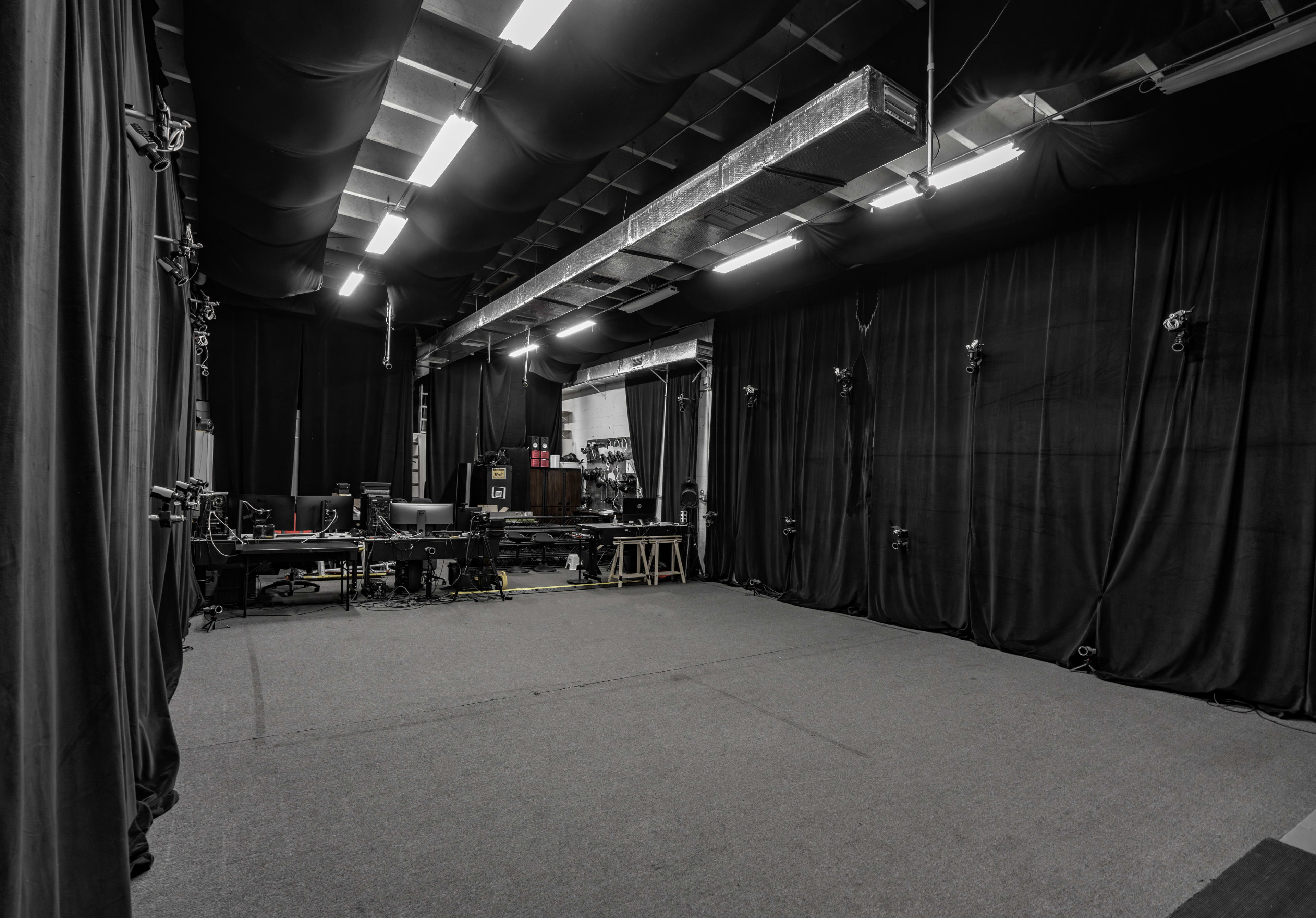 The image shows a dimly lit studio space with black curtains lining the walls, desks with equipment on one side, and various camera setups positioned around the area.
