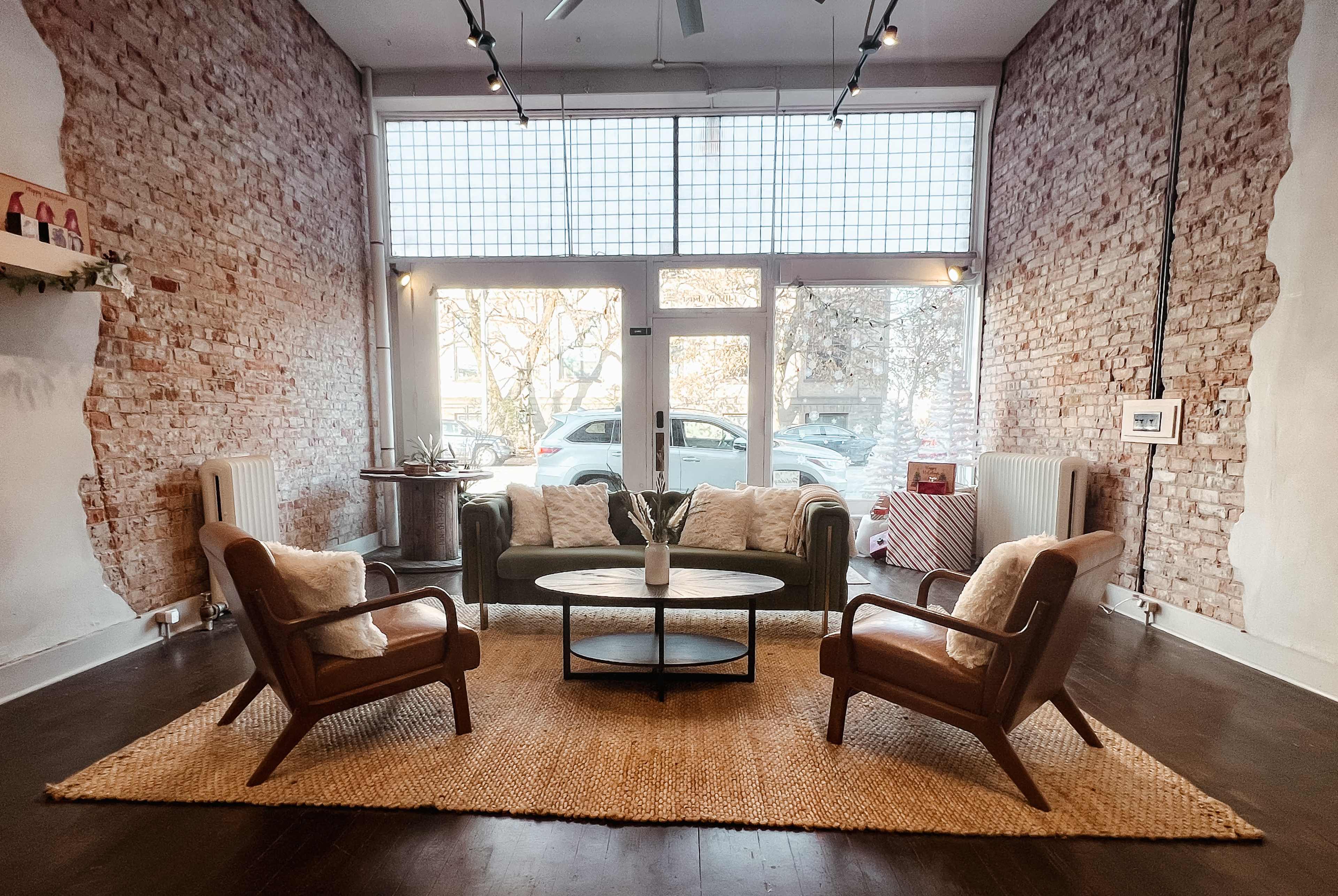 A cozy living room with exposed brick walls, a green sofa, two wooden chairs, a round coffee table, and a woven rug.