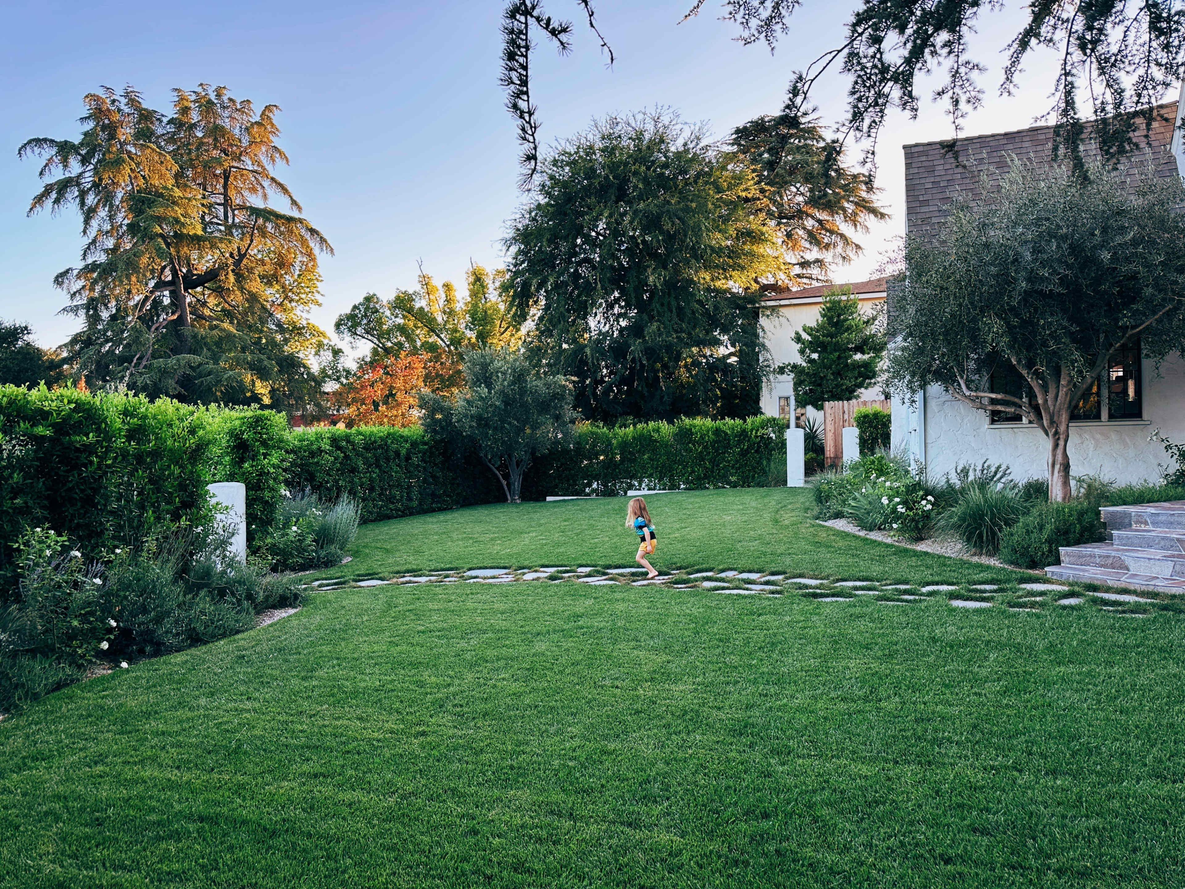 A child runs along a stone path in a landscaped yard surrounded by greenery and trees.
