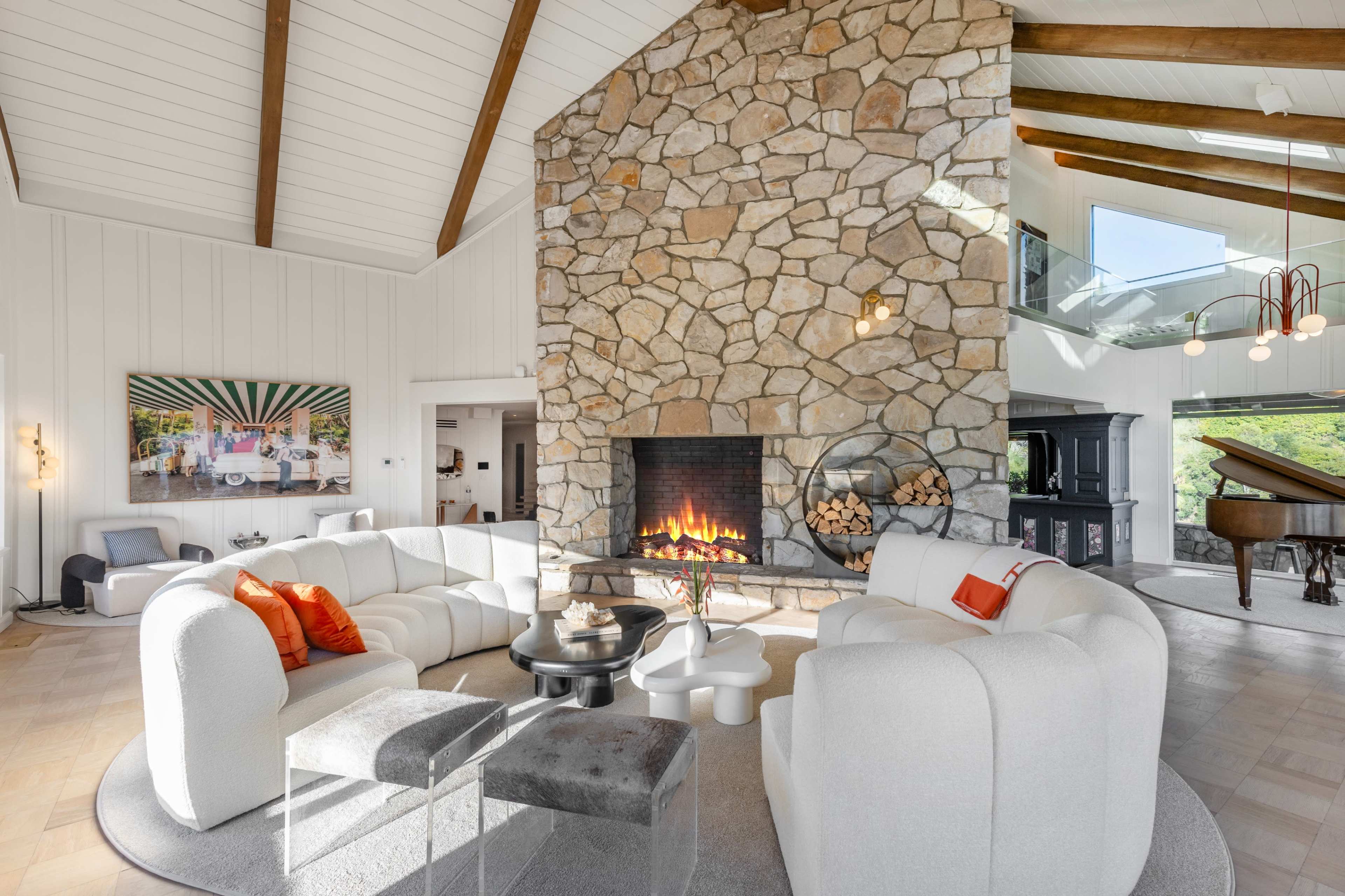 The image shows a modern living room with a stone fireplace, circular white sofas, and a grand piano in the background.