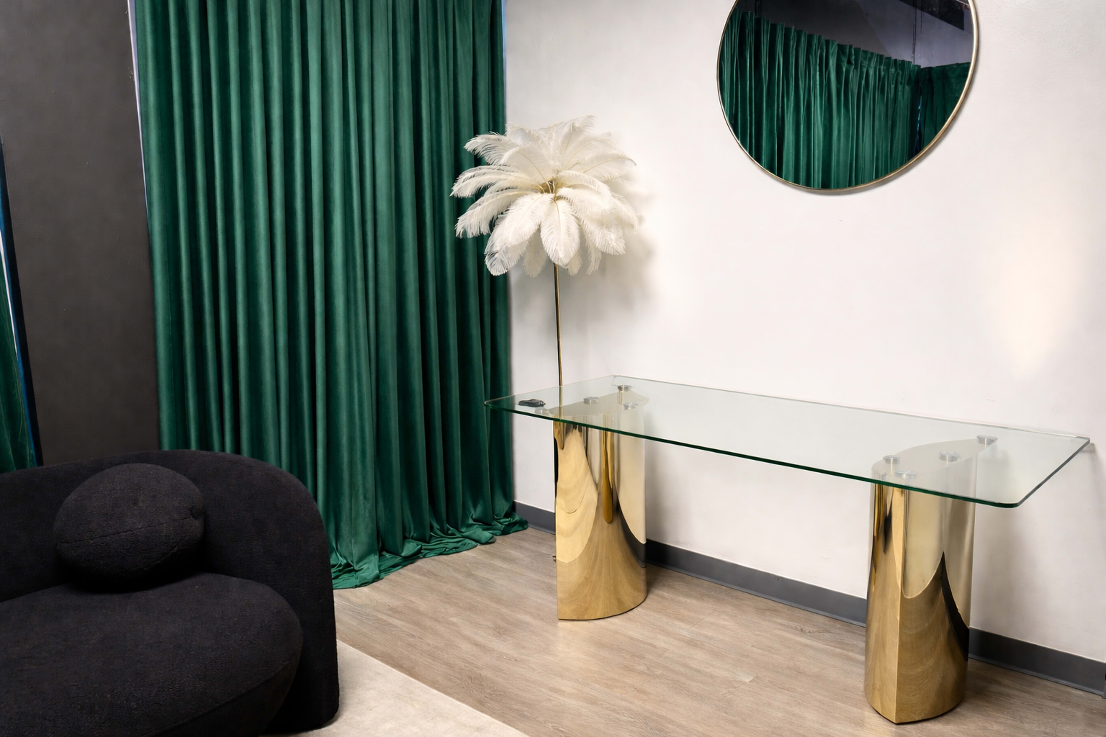 A glass console table supported by gold pillars stands against a backdrop of green curtains and a round mirror, with a decorative white feather plant nearby.