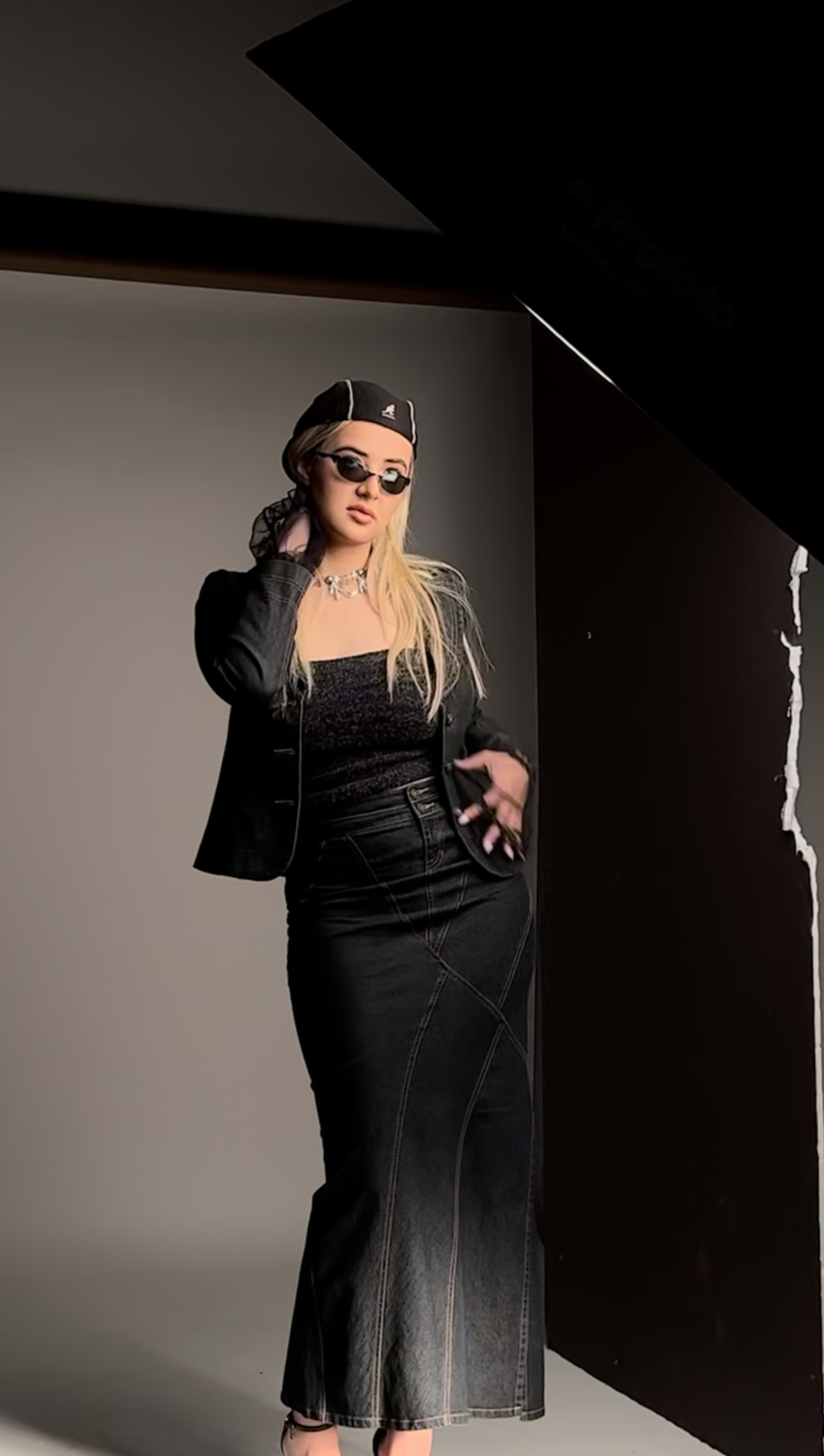 A model poses confidently in a black denim outfit with sunglasses and a headscarf in a studio setting.