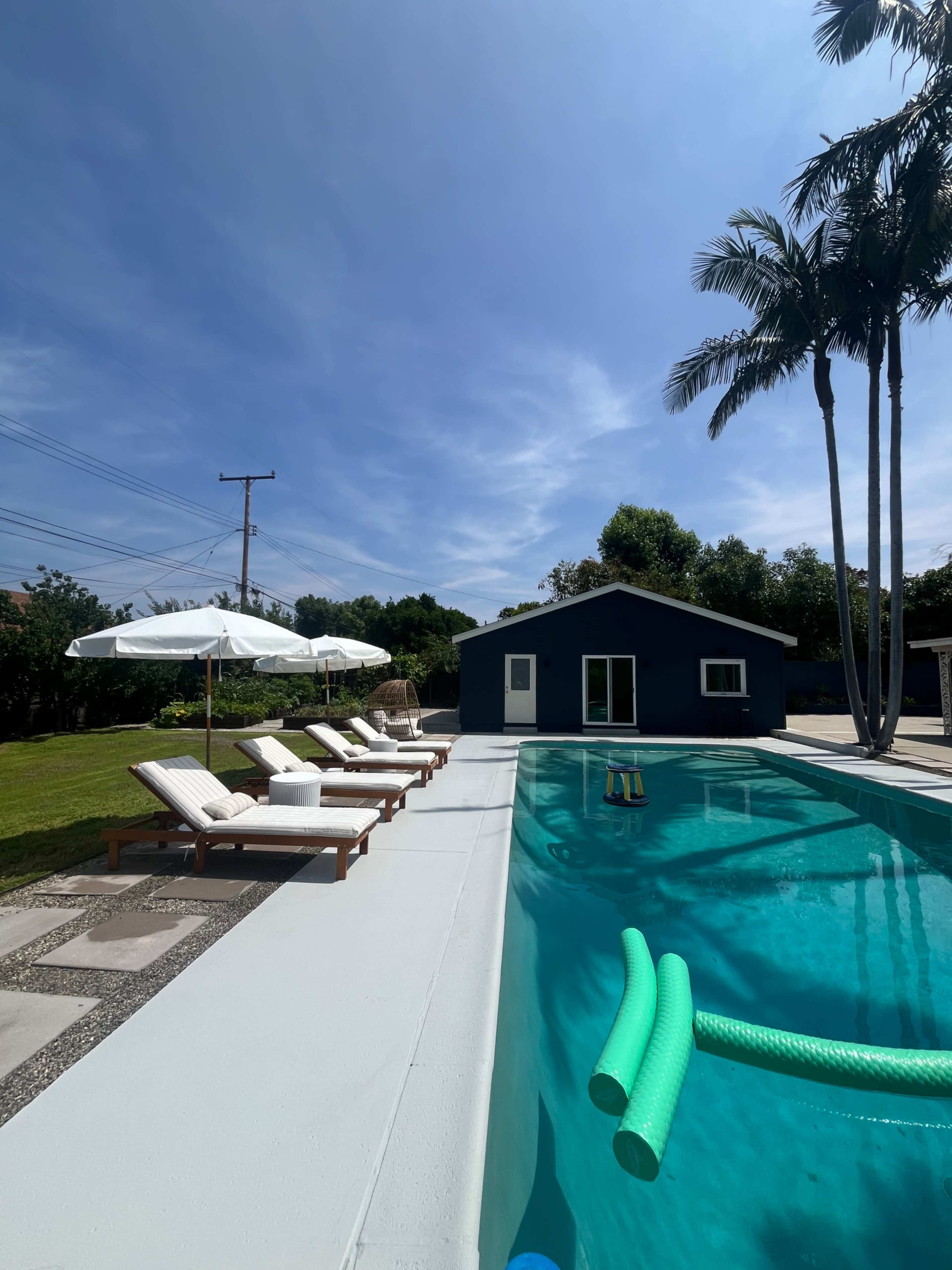 The image shows a swimming pool bordered by lounge chairs and an outdoor umbrella, with a house and palm trees visible in the background under a clear blue sky.