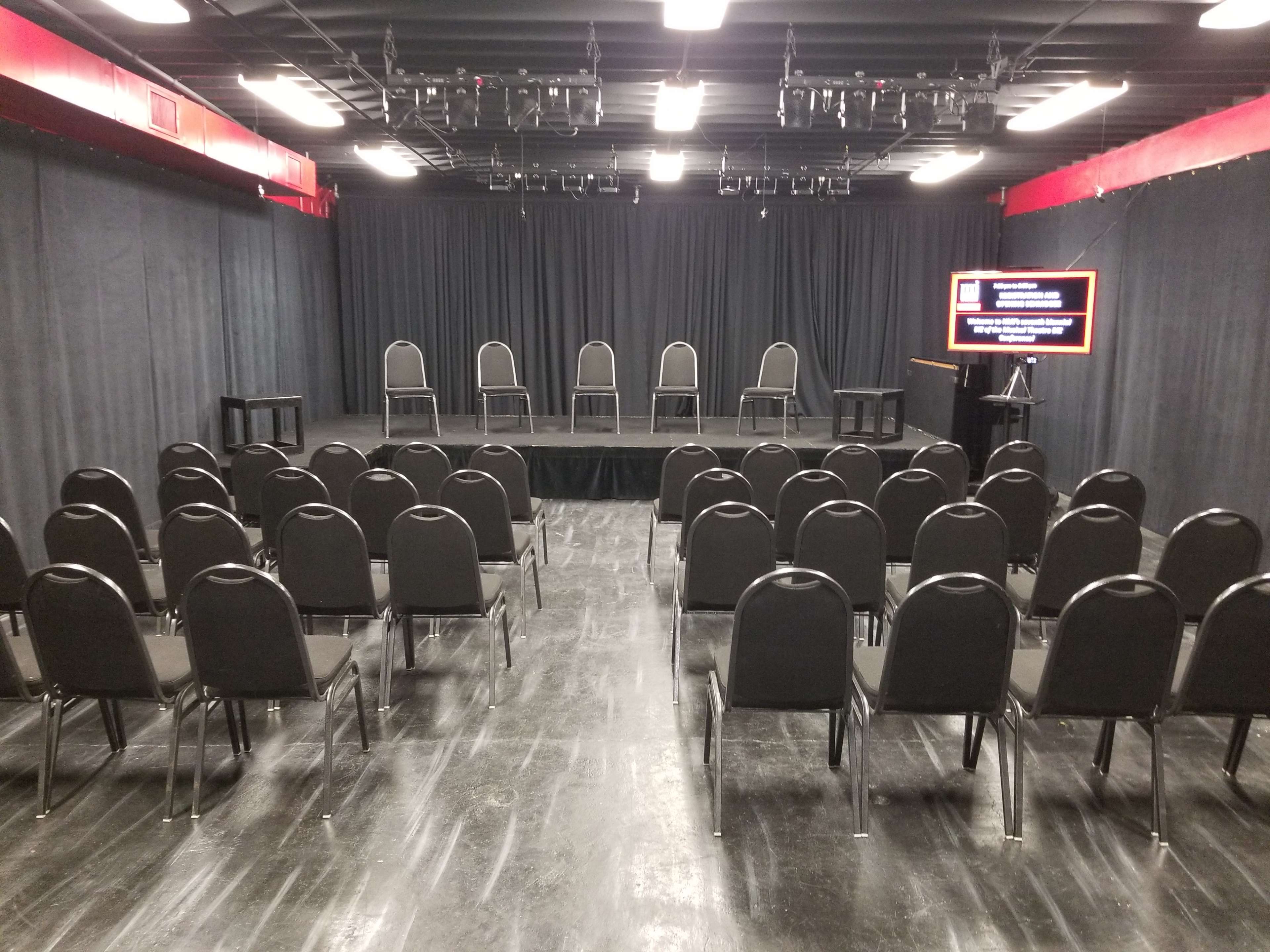 A vacant small performance space with rows of black chairs facing a short stage, surrounded by black curtains and equipped with a monitor displaying information.
