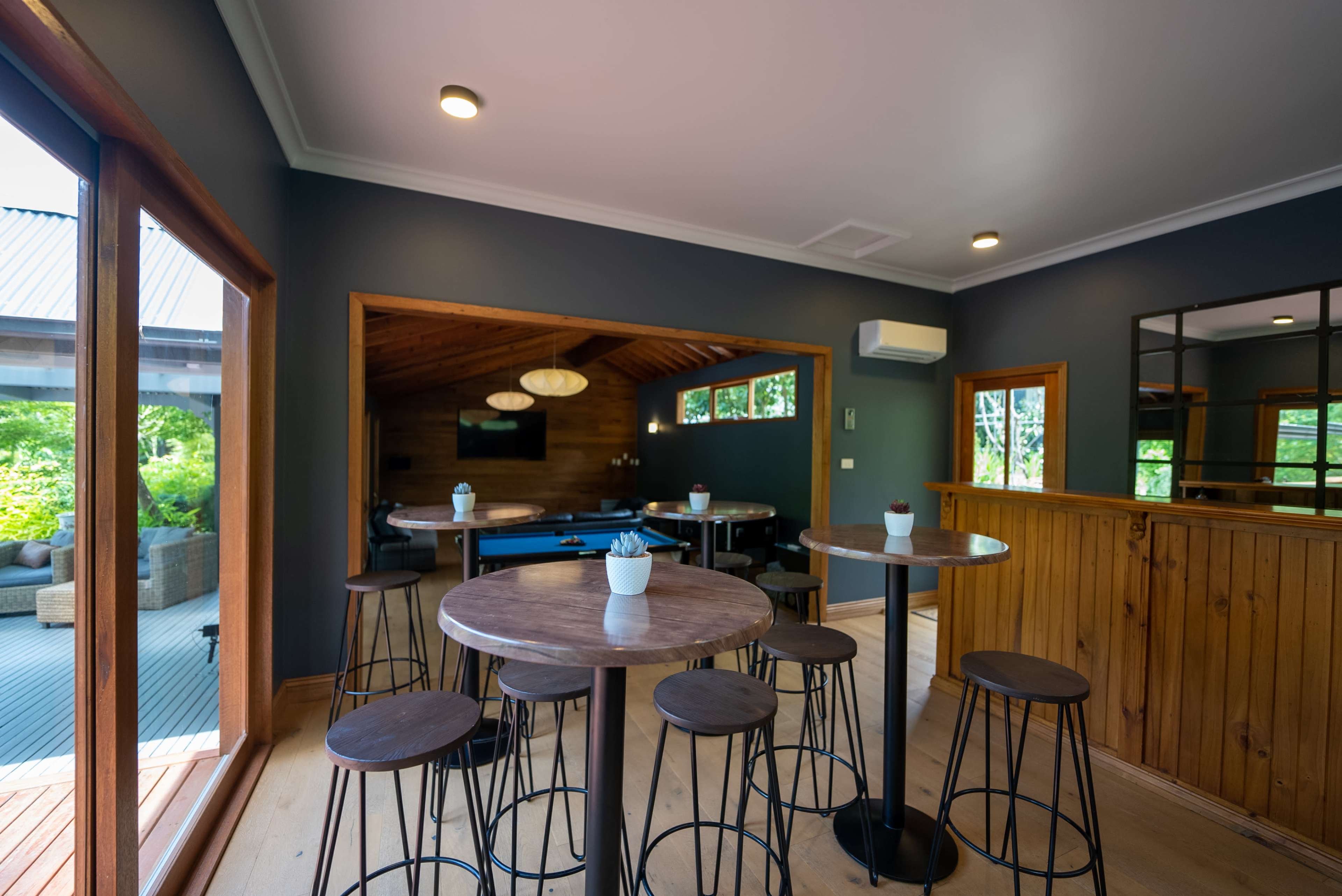 A modern interior space with several round tables and bar stools, featuring large windows overlooking a garden area.