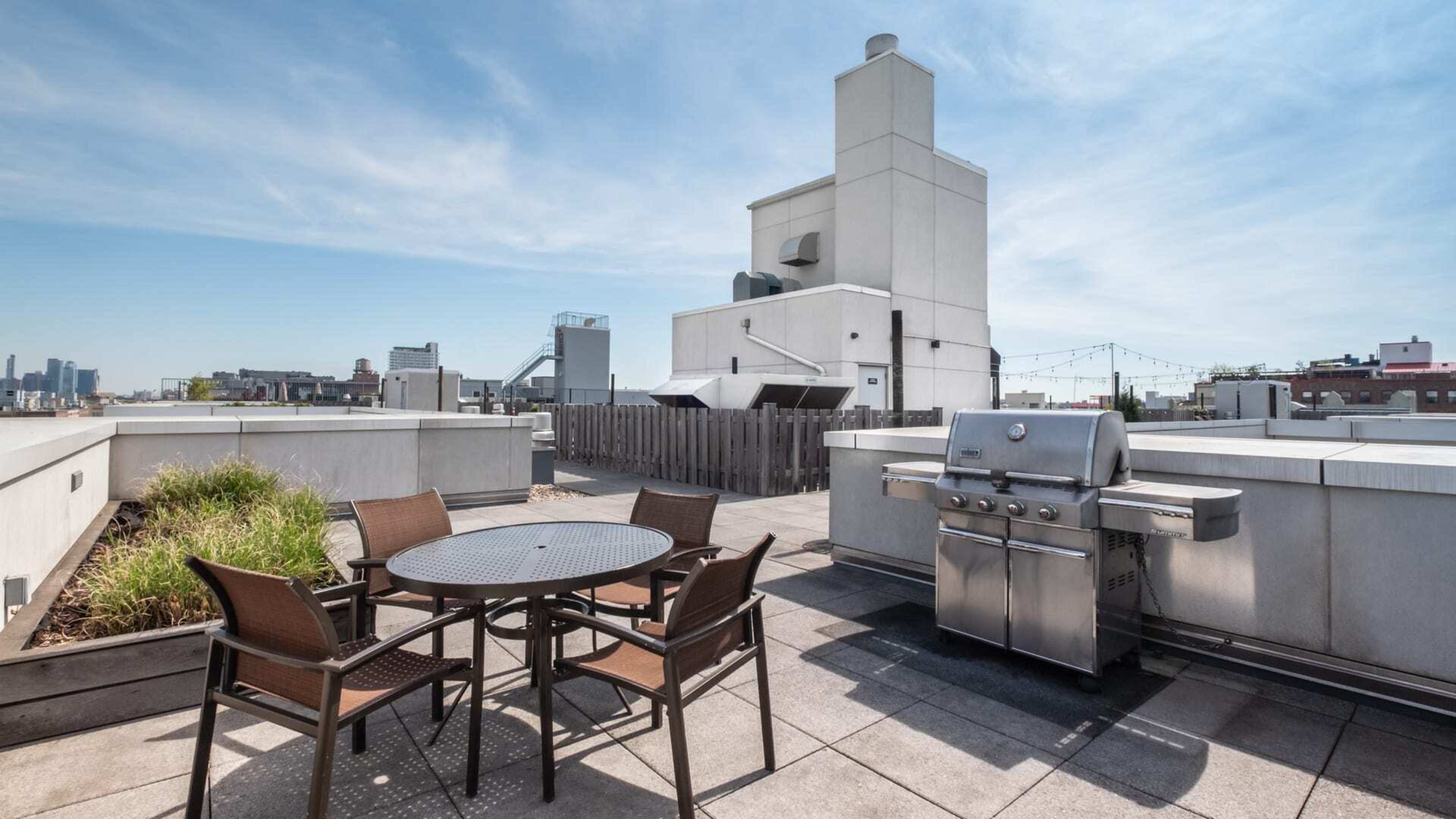 A rooftop patio featuring a circular table with four chairs and a metal grill, surrounded by urban buildings and a clear sky.