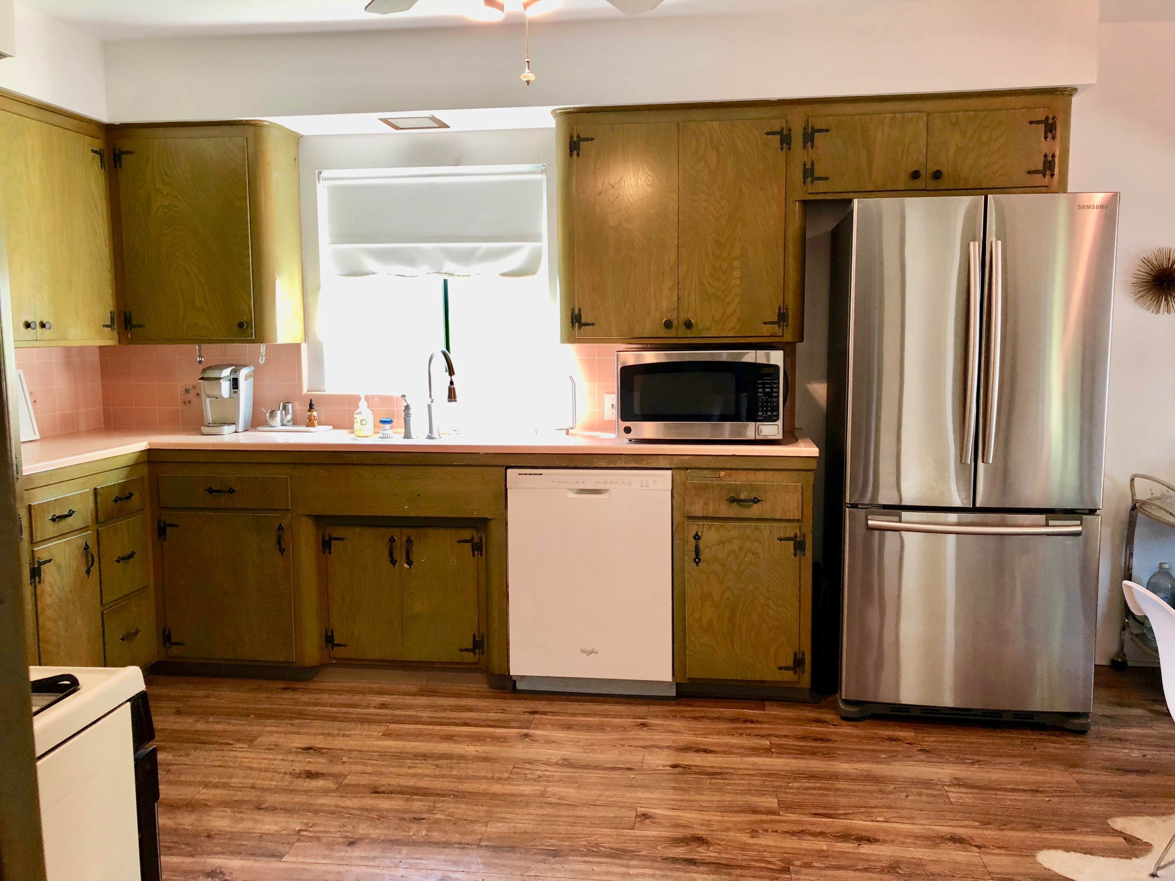 The image shows a kitchen with wooden cabinetry, a stainless steel refrigerator, a microwave, and a dishwasher, featuring a pink tile backsplash and wooden flooring.