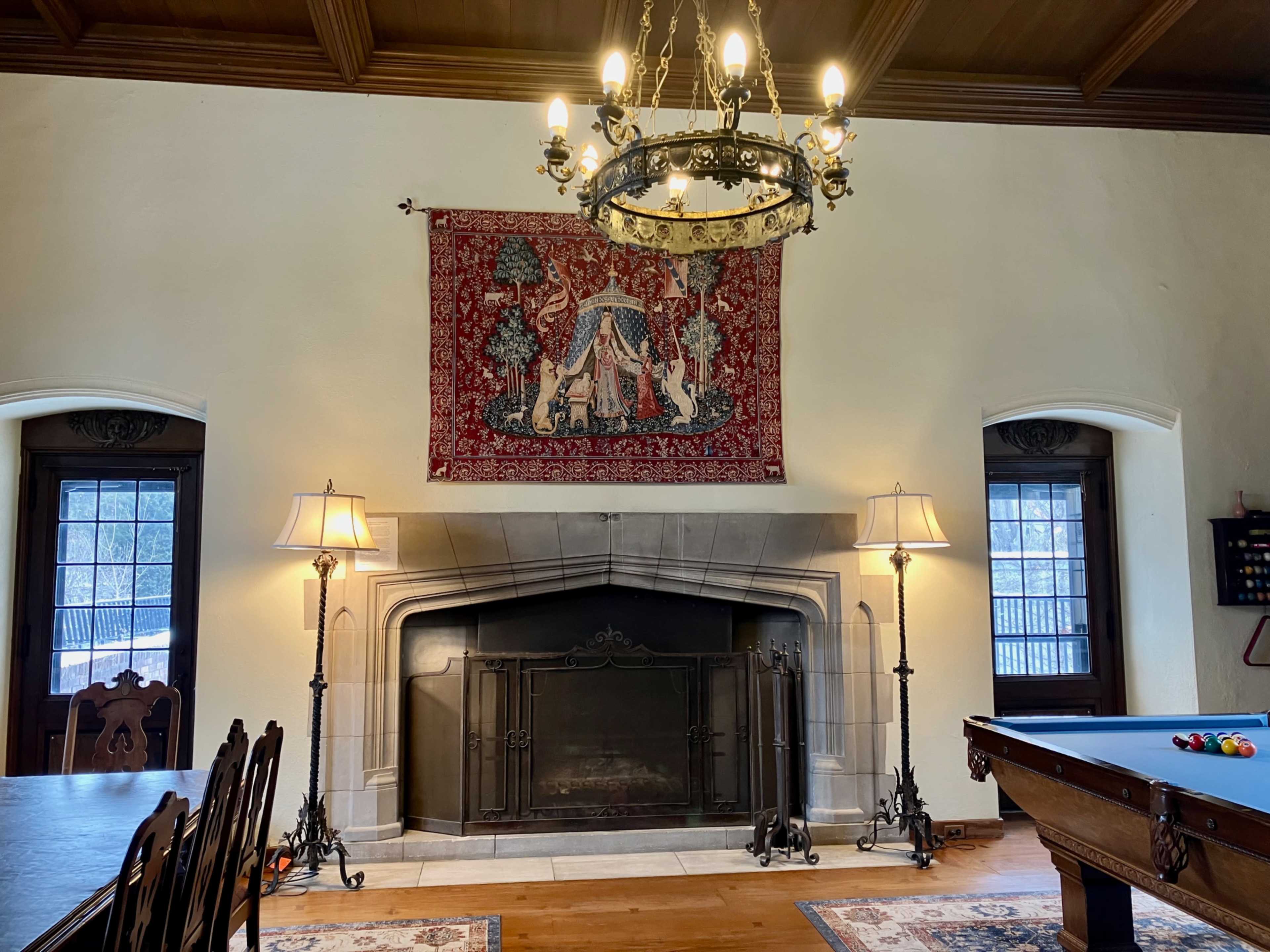 The image shows a spacious room featuring a large fireplace, a decorative tapestry on the wall, a chandelier with multiple lights, and a billiard table.
