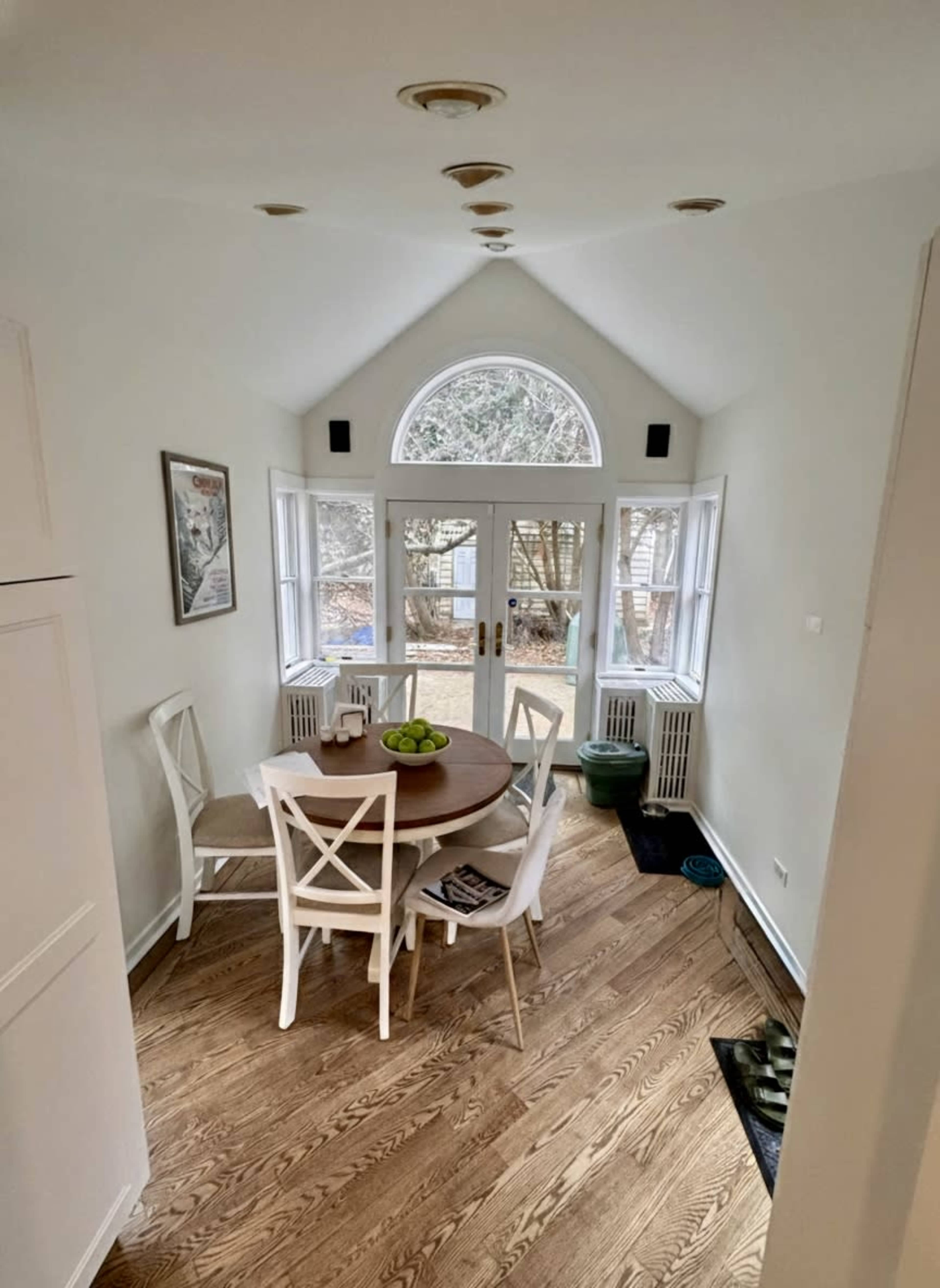 A dining area features a round table with a bowl of green apples and six white chairs, surrounded by large windows letting in natural light.