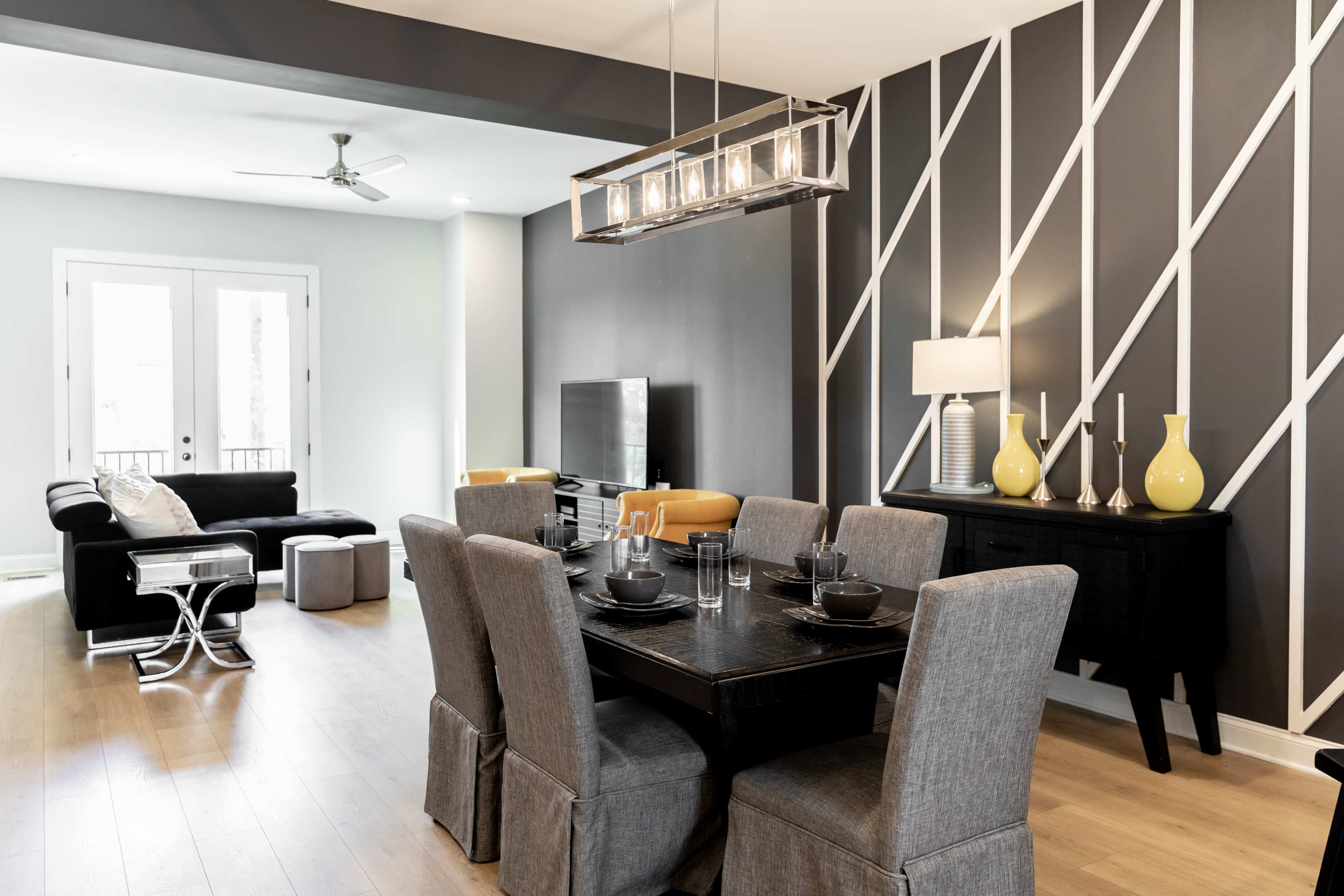 A modern dining area features a black table set for multiple diners, surrounded by gray upholstered chairs, with a living area visible in the background.