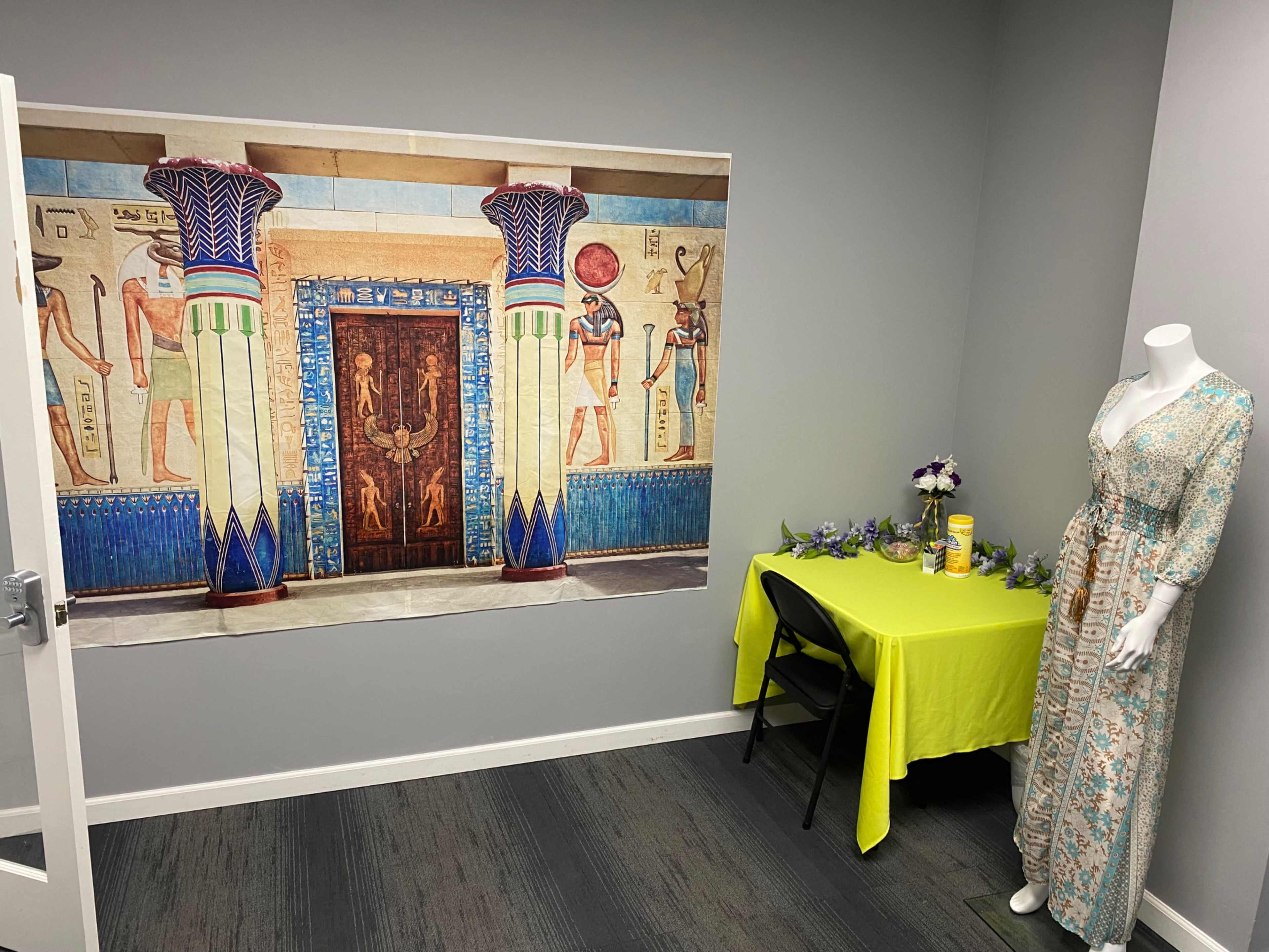 The image shows a room with a large mural of an ancient Egyptian entrance on one wall, a table covered with a bright yellow cloth, and a mannequin dressed in a patterned gown on the right.