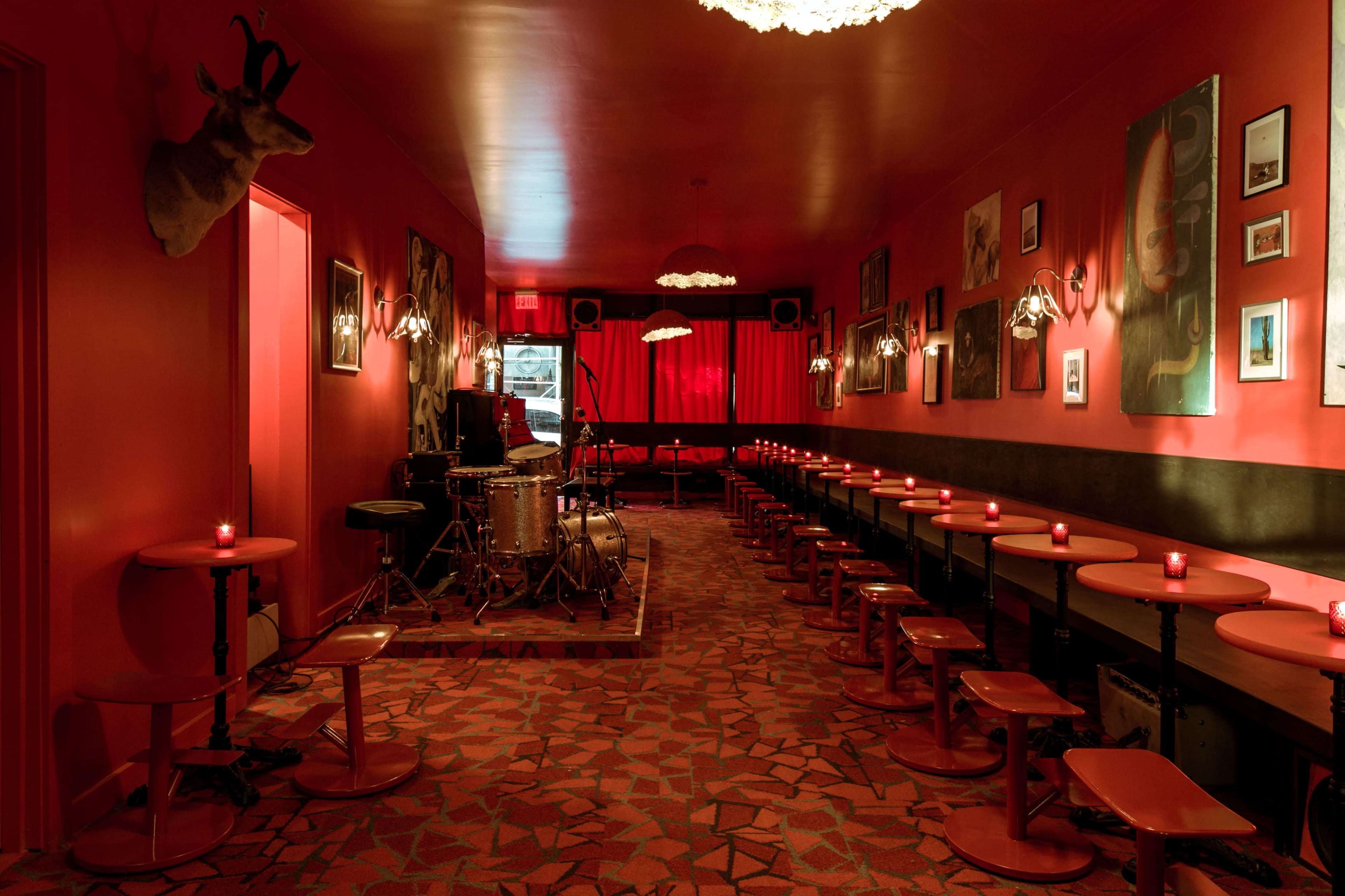 A dimly lit lounge with red walls, circular tables, and an arrangement of chairs, featuring a small stage for live music at one end.