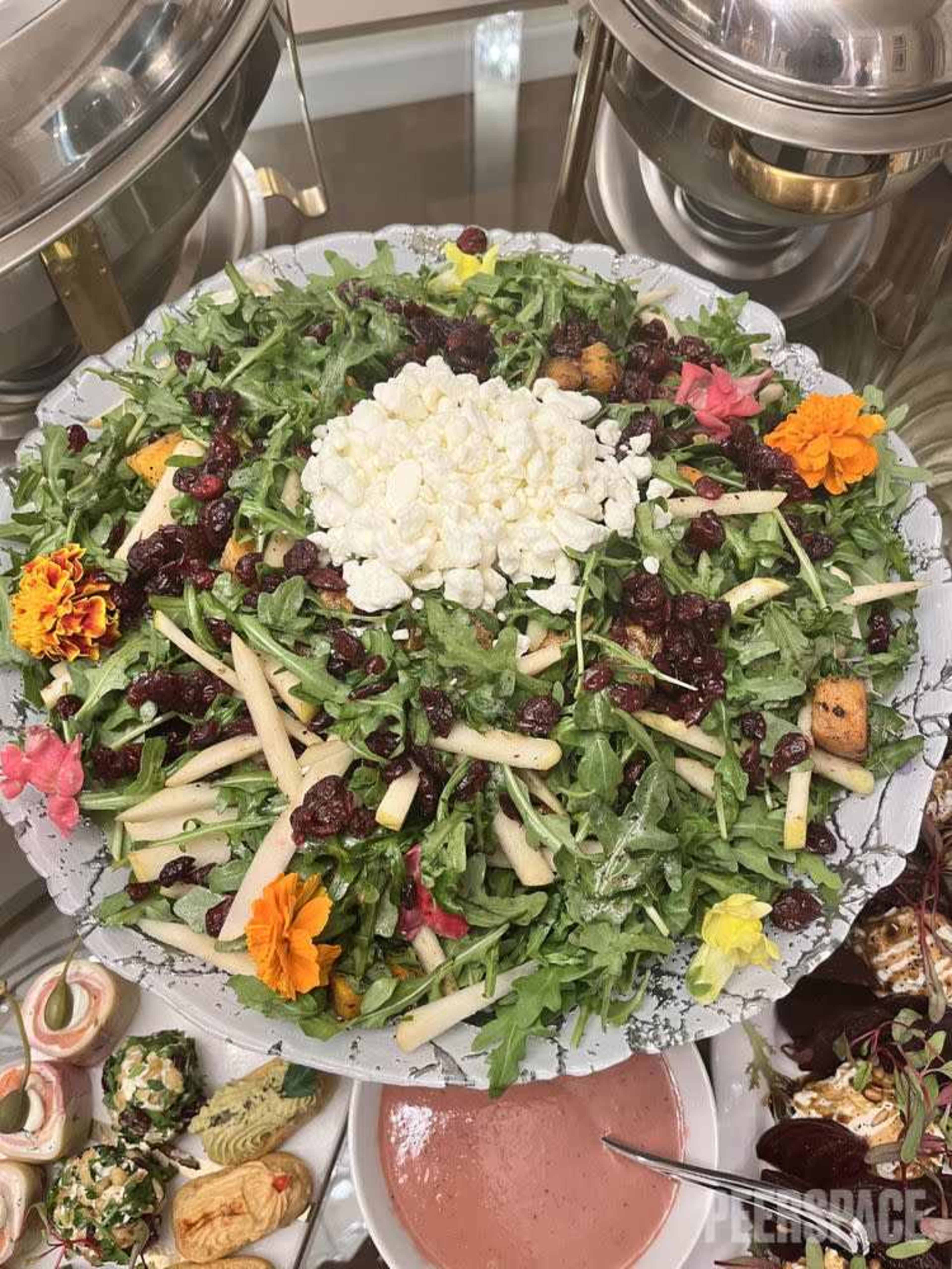 A large platter of salad featuring arugula, dried cranberries, apples, goat cheese, and edible flowers.