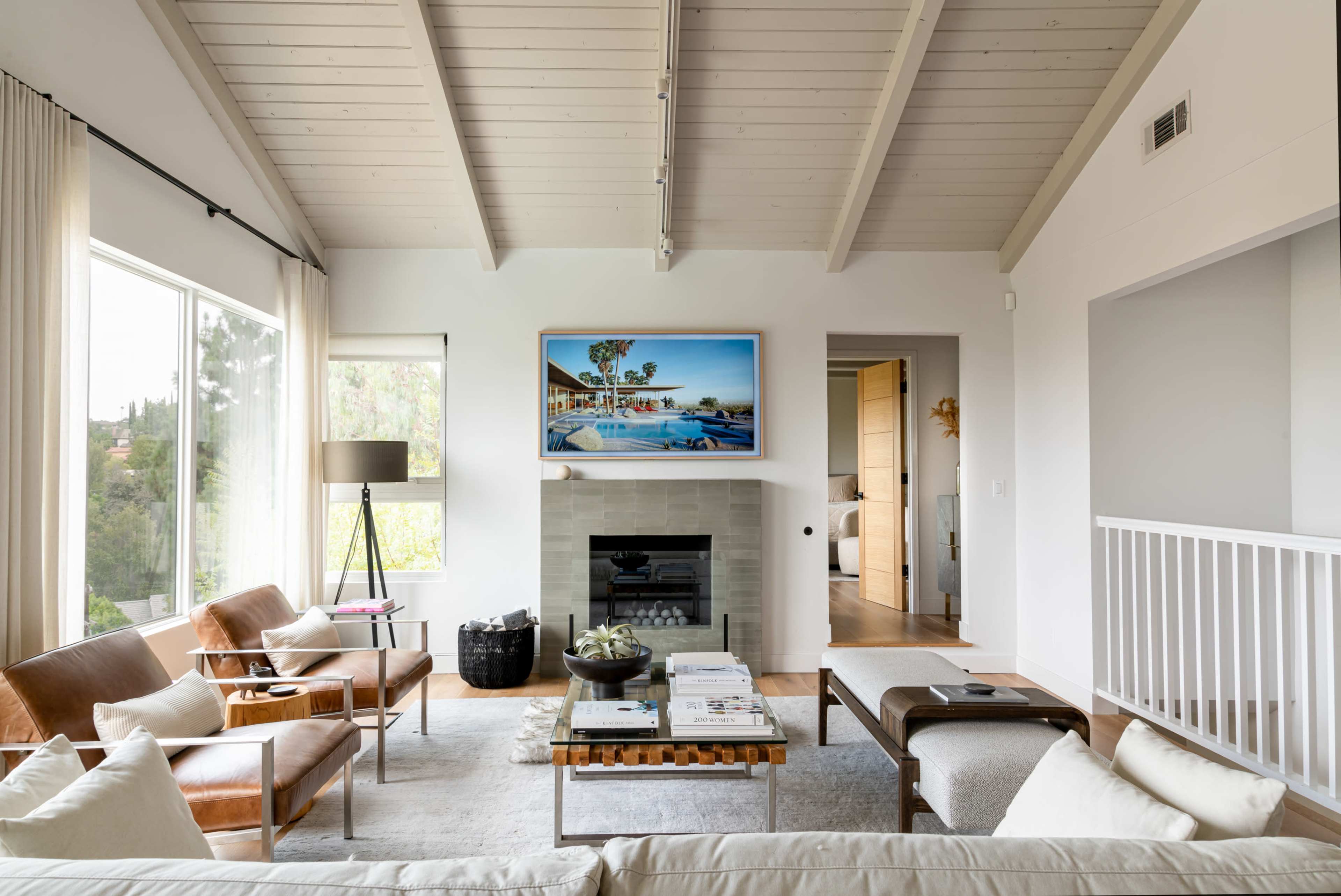 A modern living room with a fireplace, large windows, and a couch, featuring neutral tones and wooden ceiling beams.