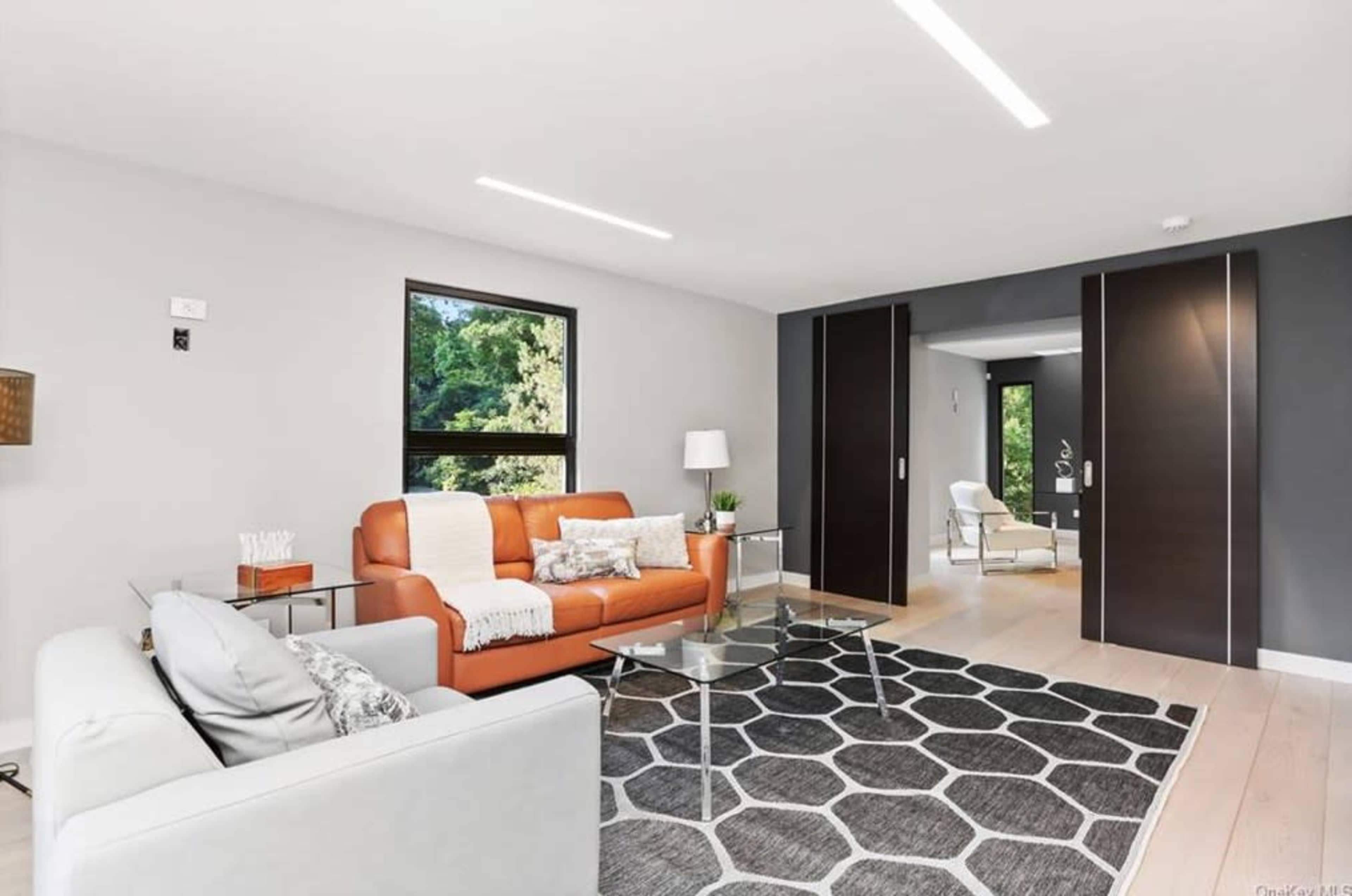 The image shows a modern living room with a brown leather sofa, a white sofa, a glass coffee table, and large windows overlooking greenery.