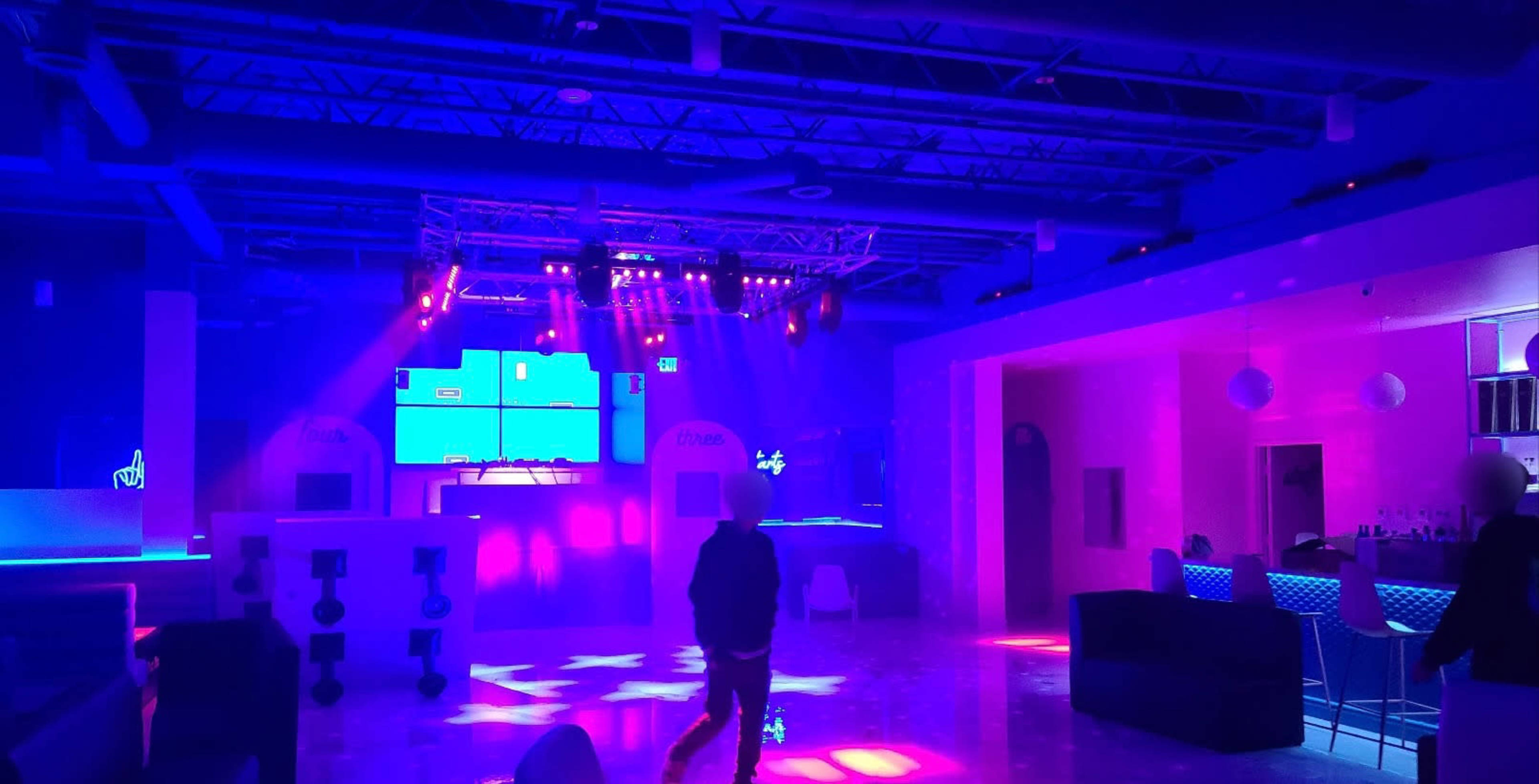 A dimly lit nightclub features purple and blue lighting with a DJ booth in the background and scattered seating areas.