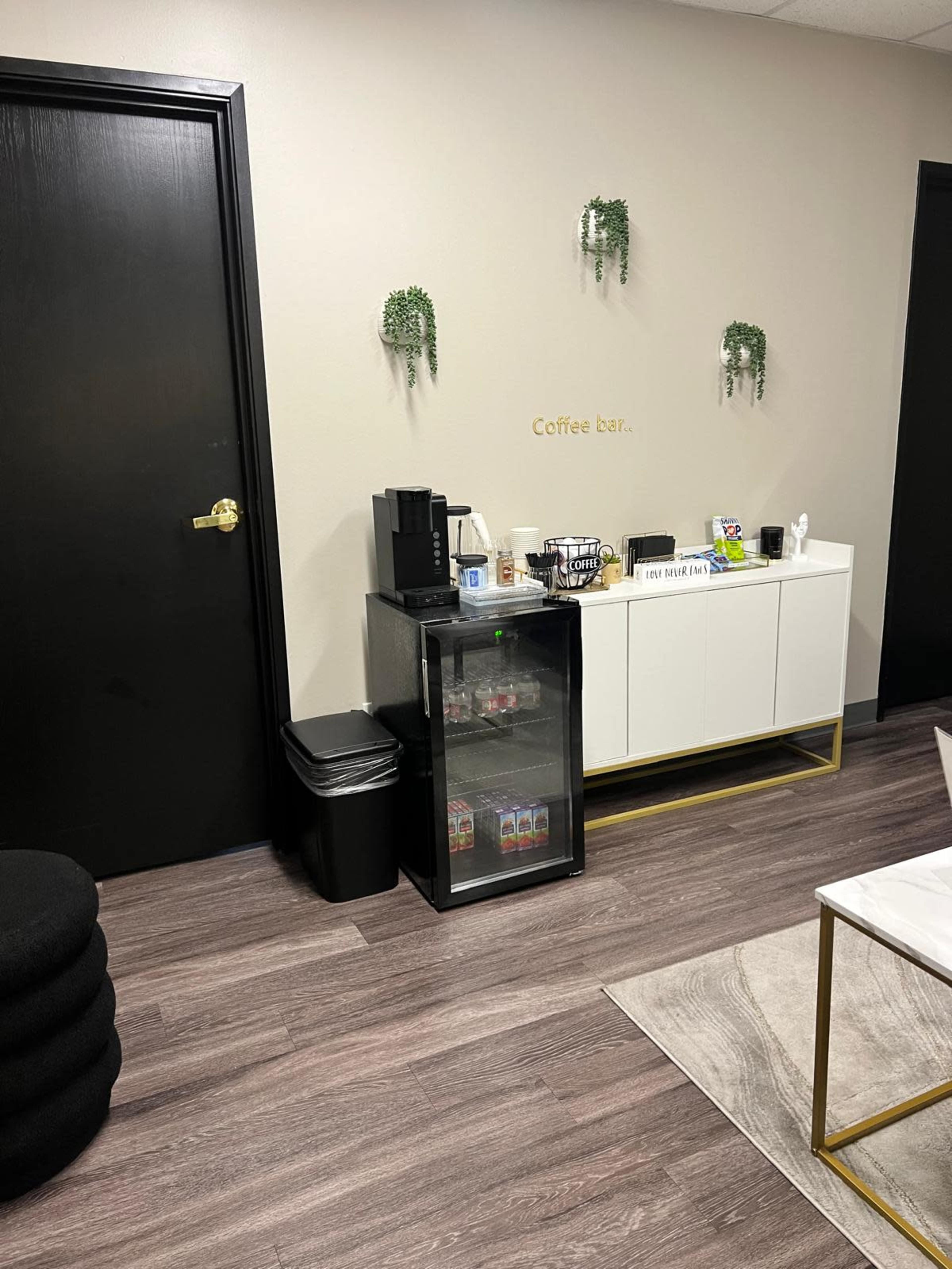 The image shows a small waiting area featuring a coffee station, a trash can, and a black cabinet under a wall with green plants.
