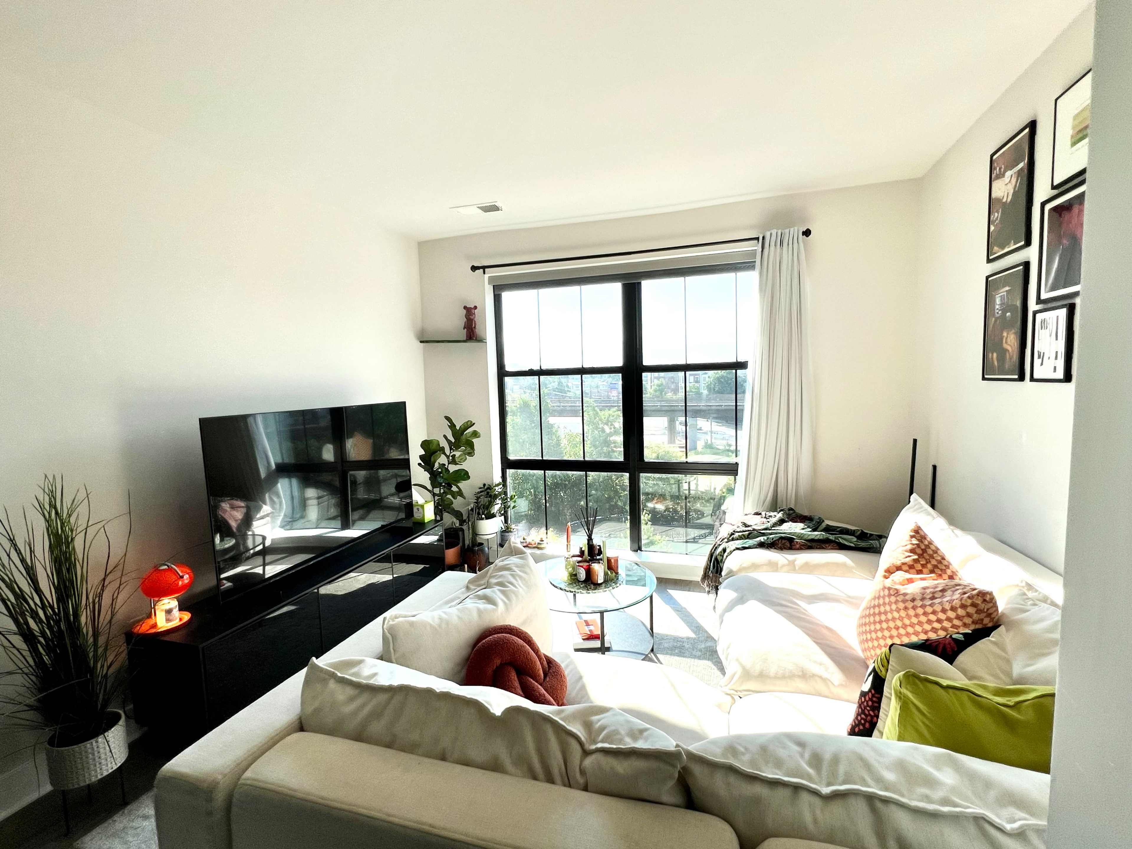 A bright living room features a sectional sofa, a coffee table, a large television, and a window with a view.