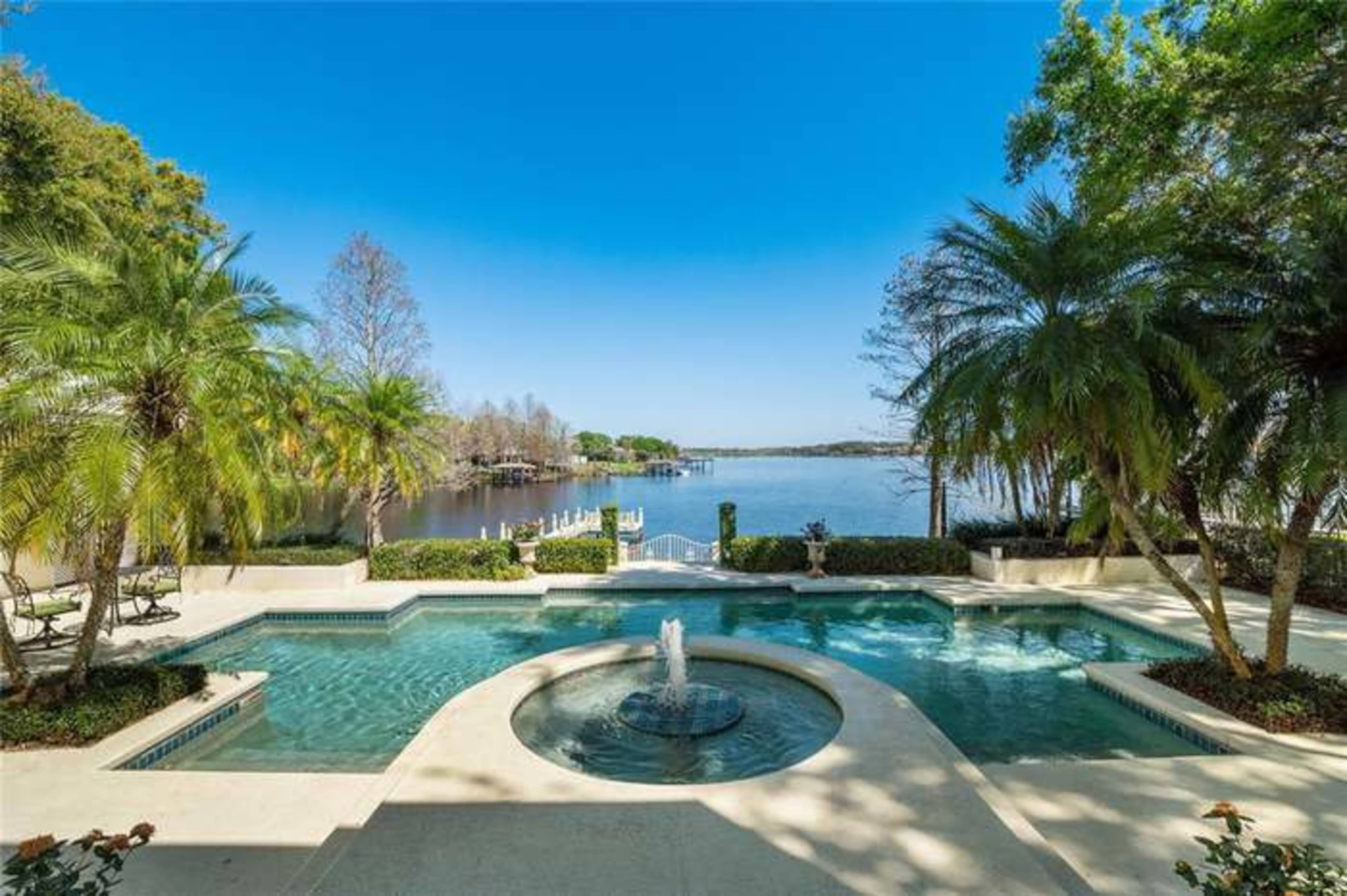 A landscaped pool area with a fountain, overlooking a calm lake surrounded by trees and greenery.