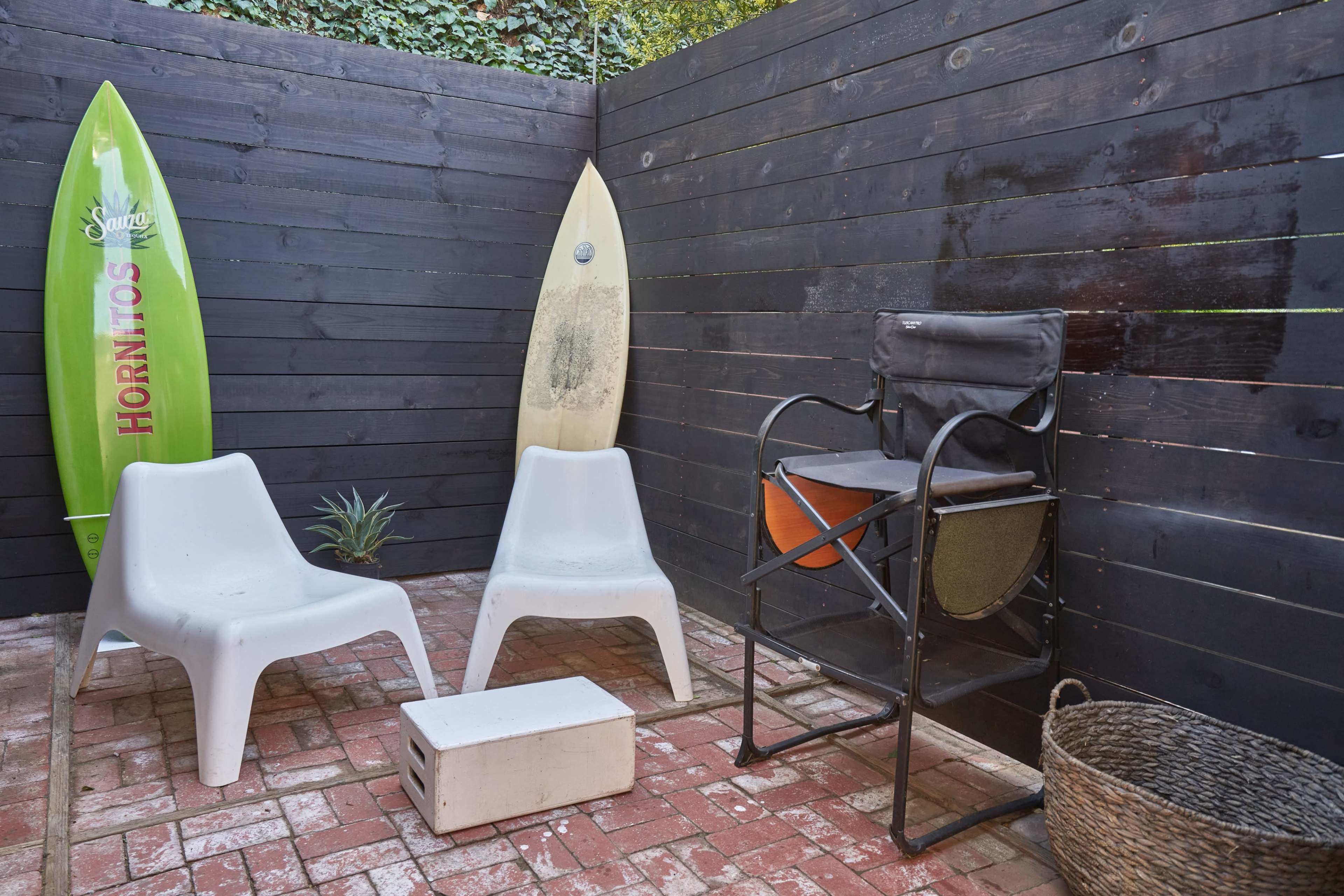 A small outdoor area with two white chairs, a folding chair, a wooden crate, and two surfboards leaning against a black wooden fence.