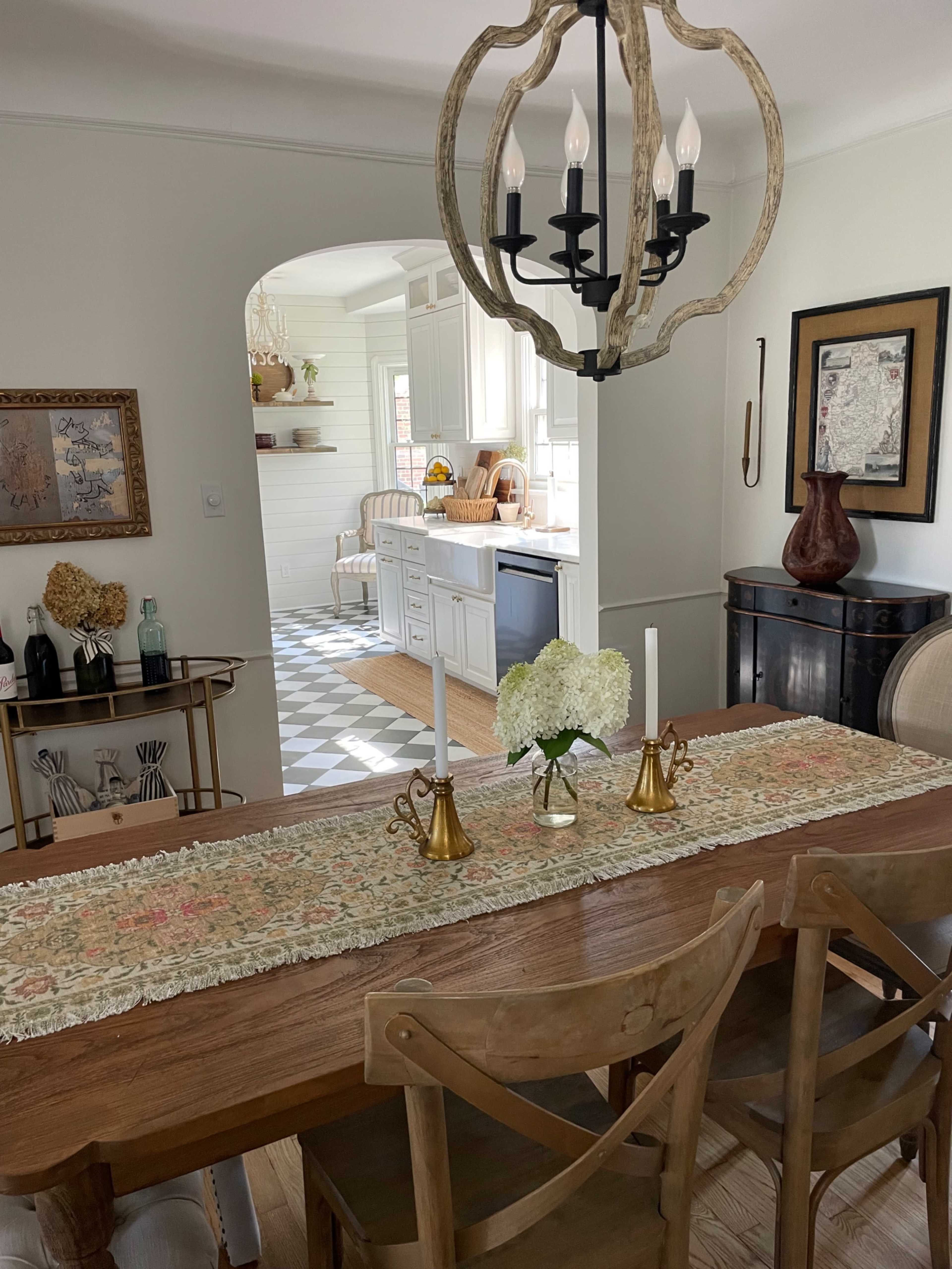 A wooden dining table is set with two candlesticks and a floral table runner, while a doorway leads to a kitchen with light cabinets and a checkered floor.