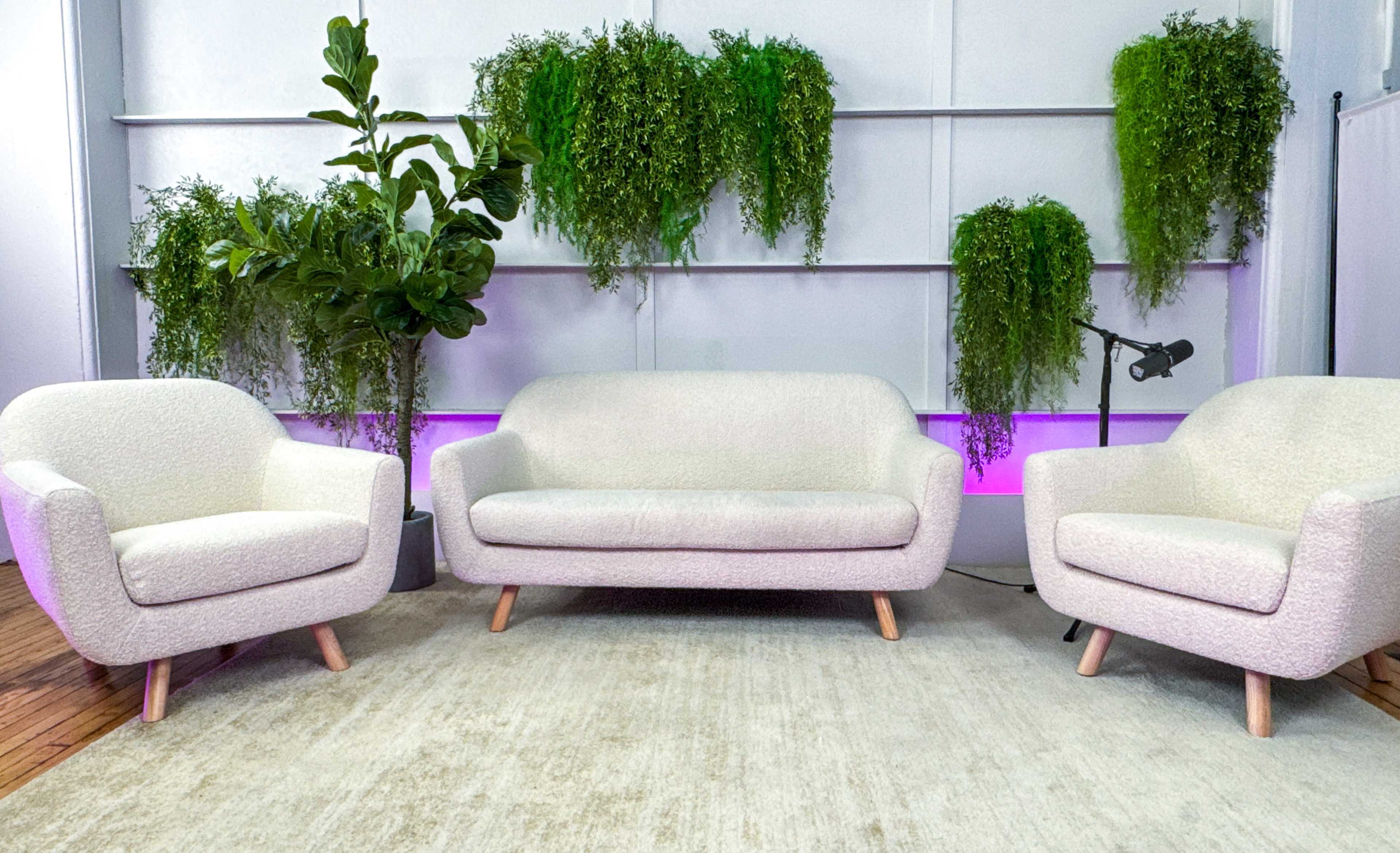 A light-colored, textured sofa and two matching armchairs are arranged on a rug in a room with green hanging plants and purple ambient lighting.