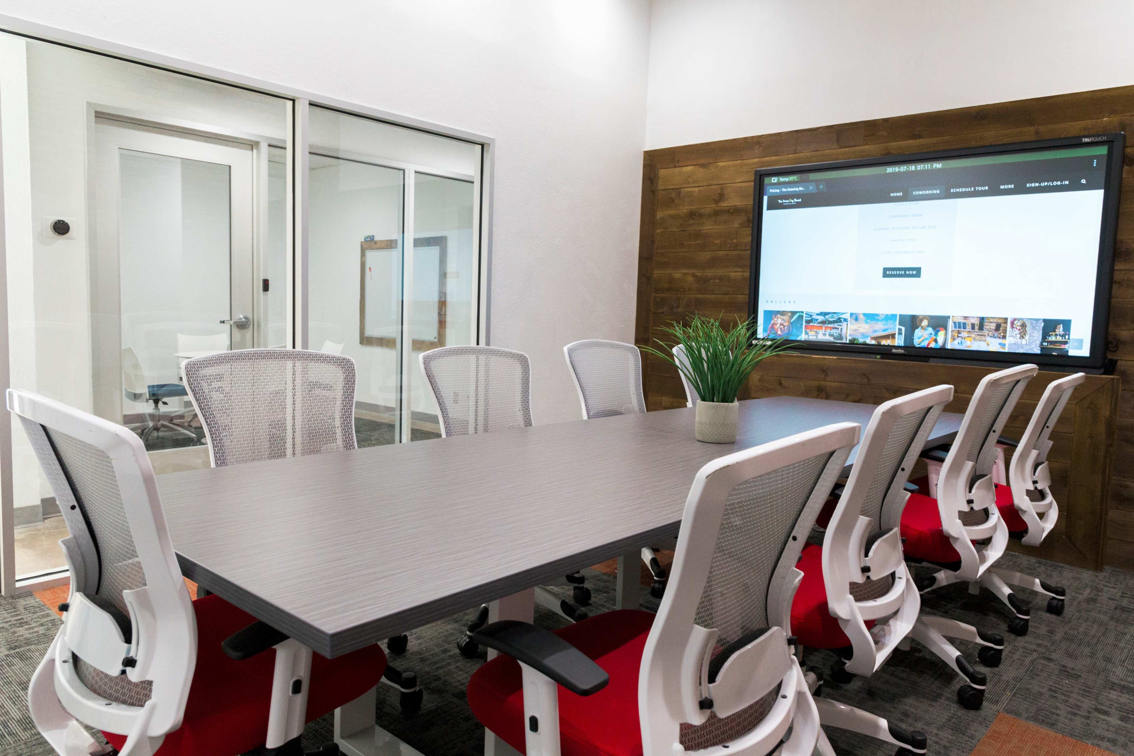 A conference room features a large table surrounded by white and red chairs, with a screen displaying a web page on the wall.