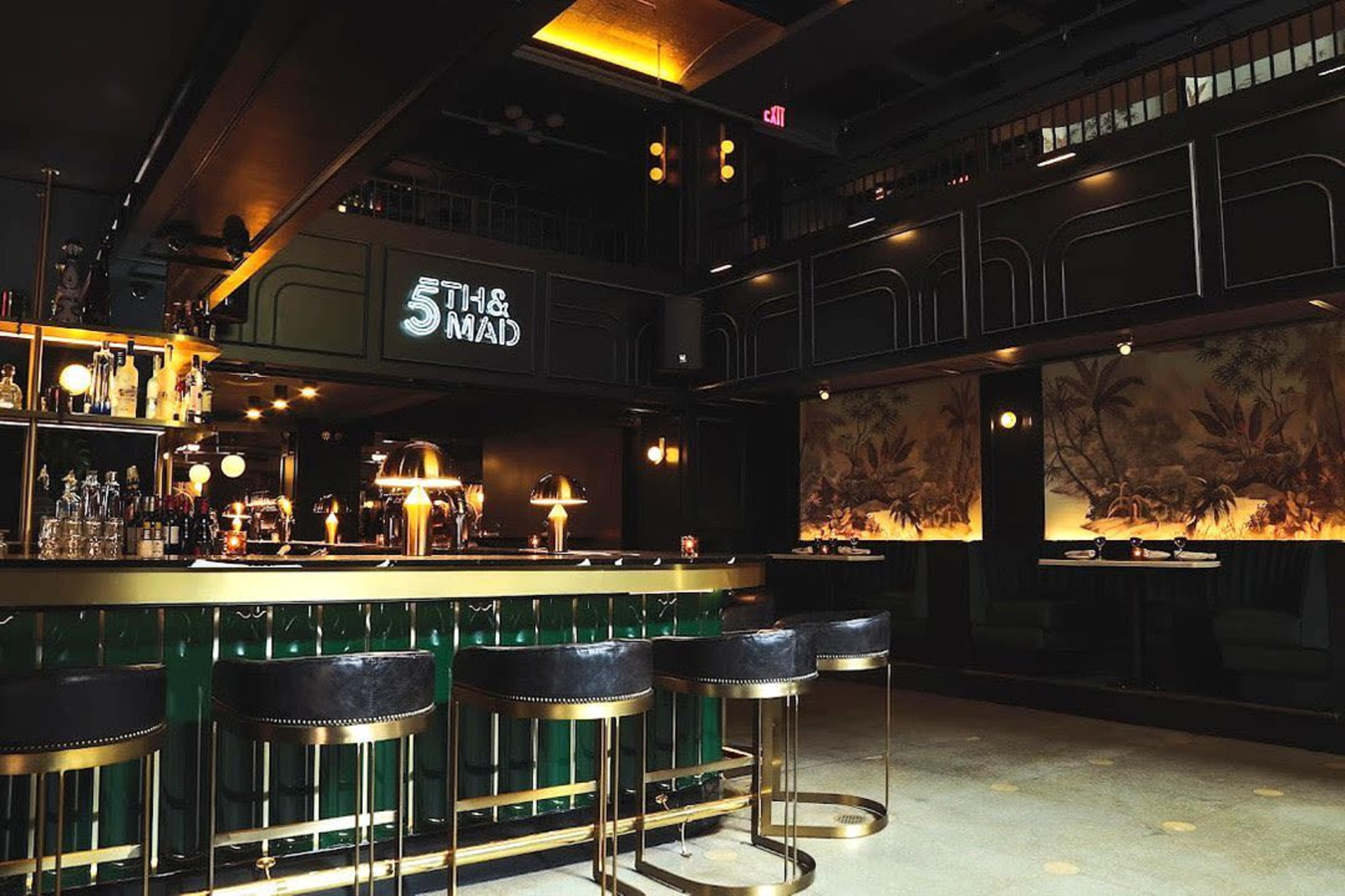 The image shows a stylish bar area with green decor, a polished counter, and artistic wall displays, illuminated by warm lighting.