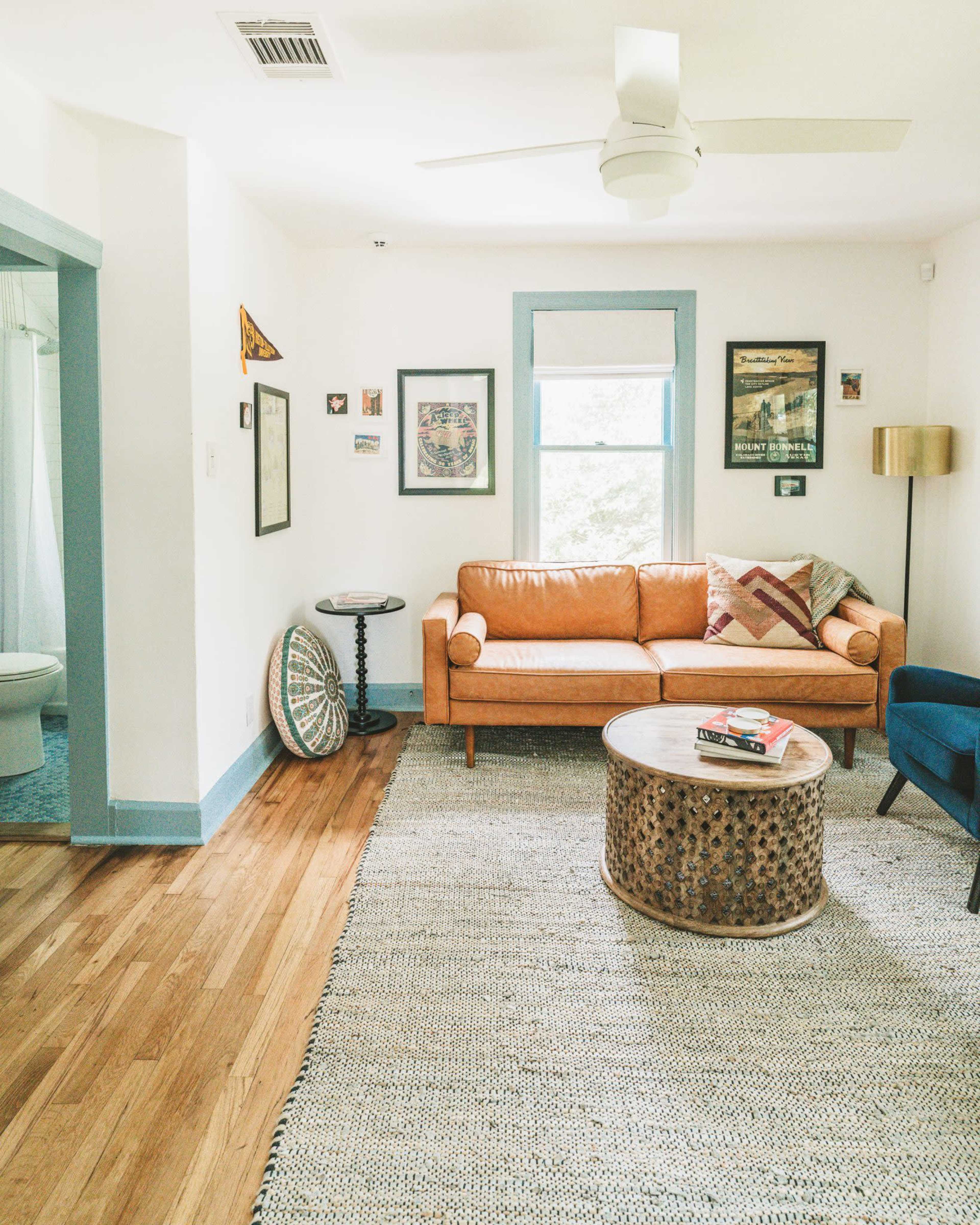 A well-lit living room features a brown leather sofa, a blue armchair, a round coffee table, and various framed artworks on the walls.