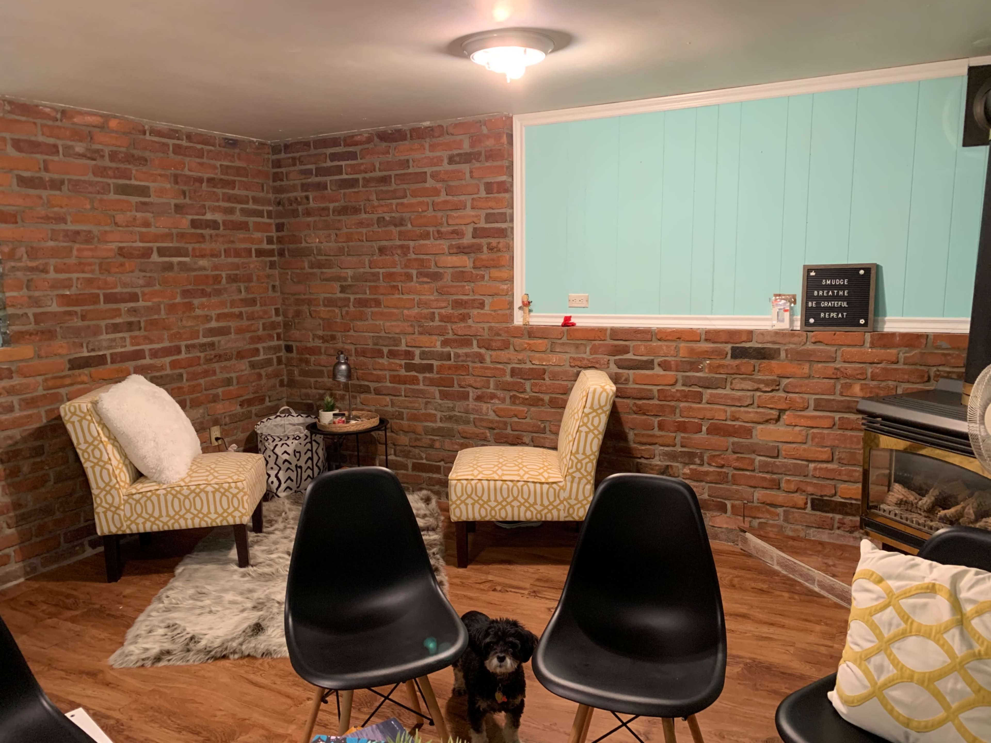 A cozy room with exposed brick walls, two patterned chairs, and a dog on a wooden floor, accompanied by a fireplace and a small table.