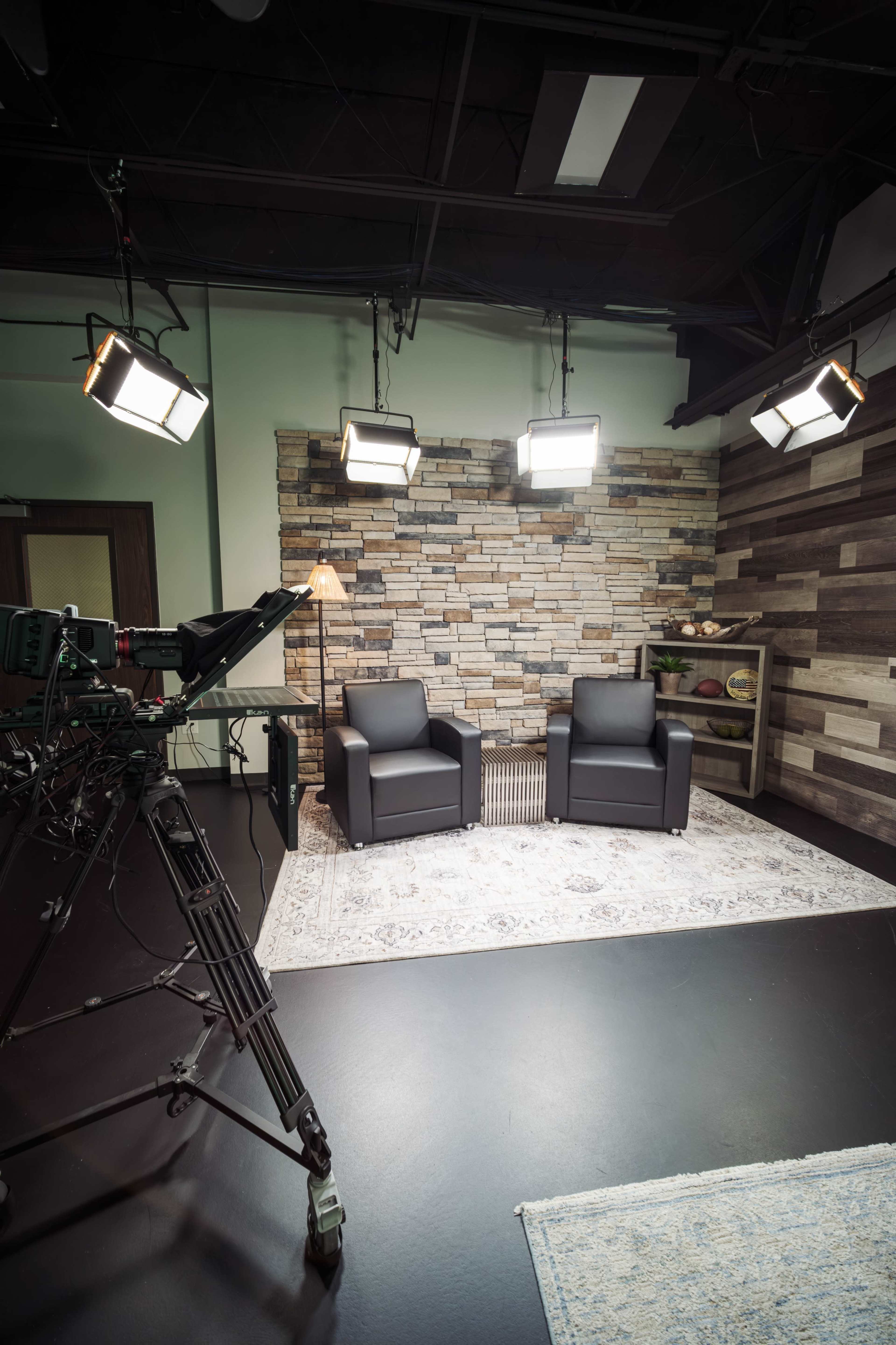 The image shows a TV studio setup with two black chairs facing a stone wall, illuminated by overhead lights, and a camera positioned nearby.