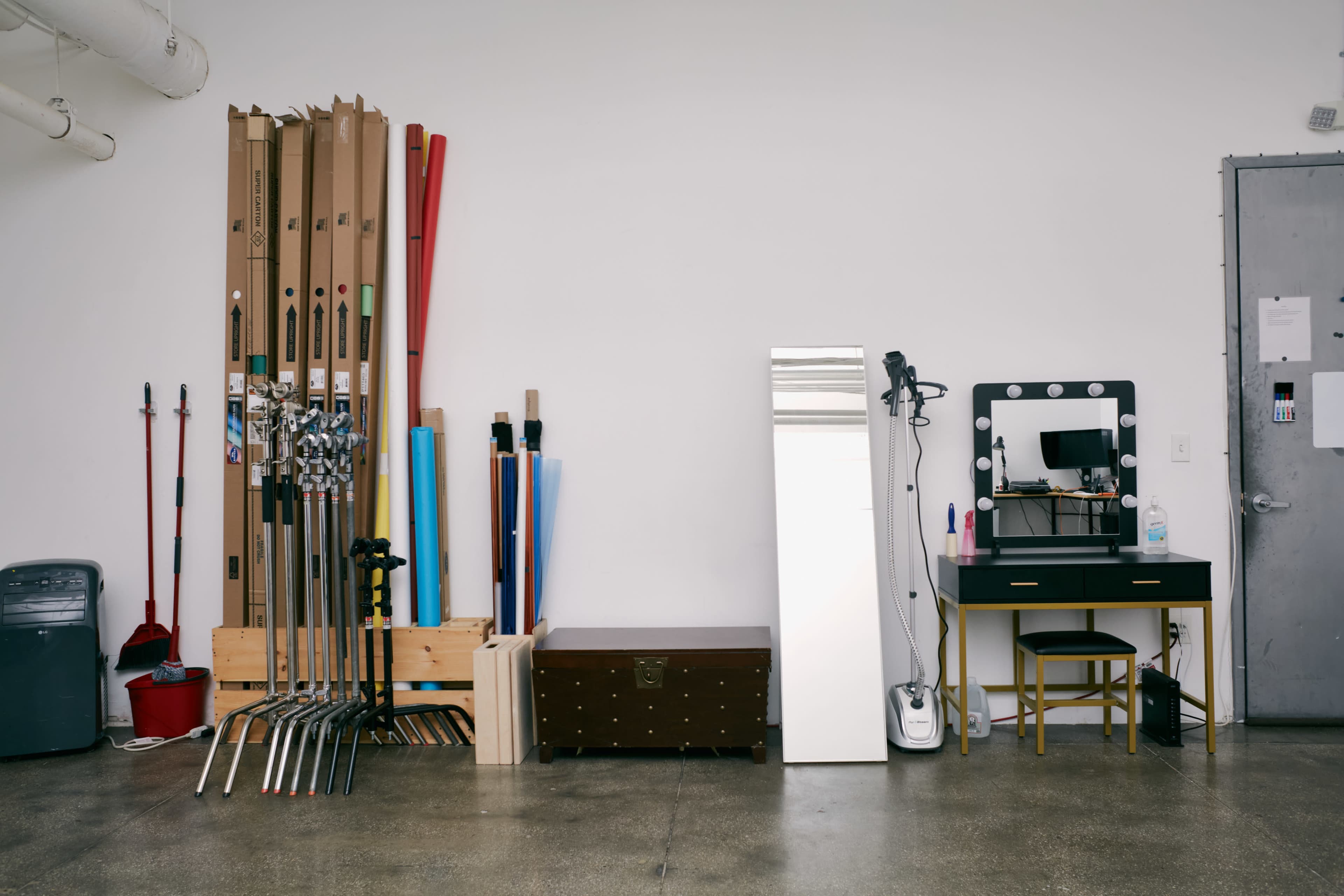 The image shows a workspace with a variety of tools and materials, including cardboard tubes, garden tools, a mirror, a vanity table, and a storage chest against a plain wall.