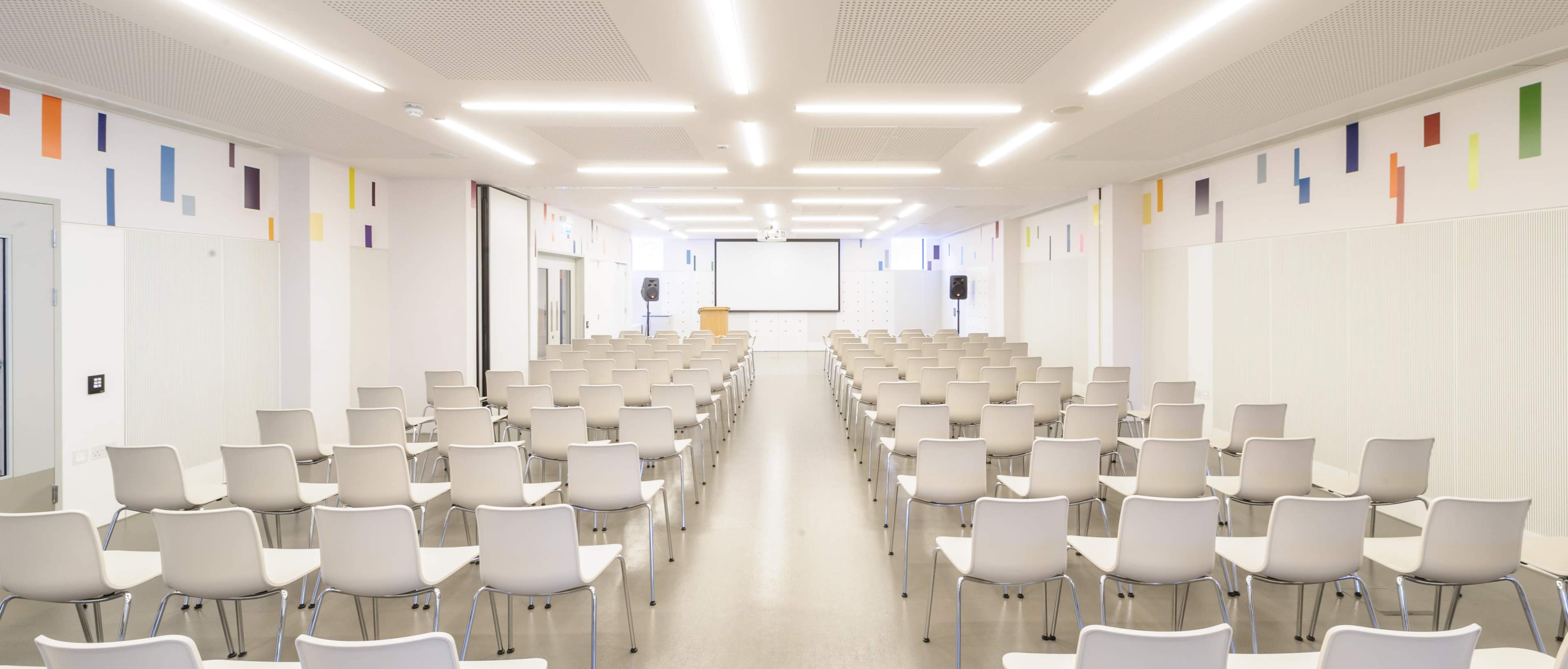 The image shows a large, modern conference room filled with rows of empty chairs facing a projection screen.