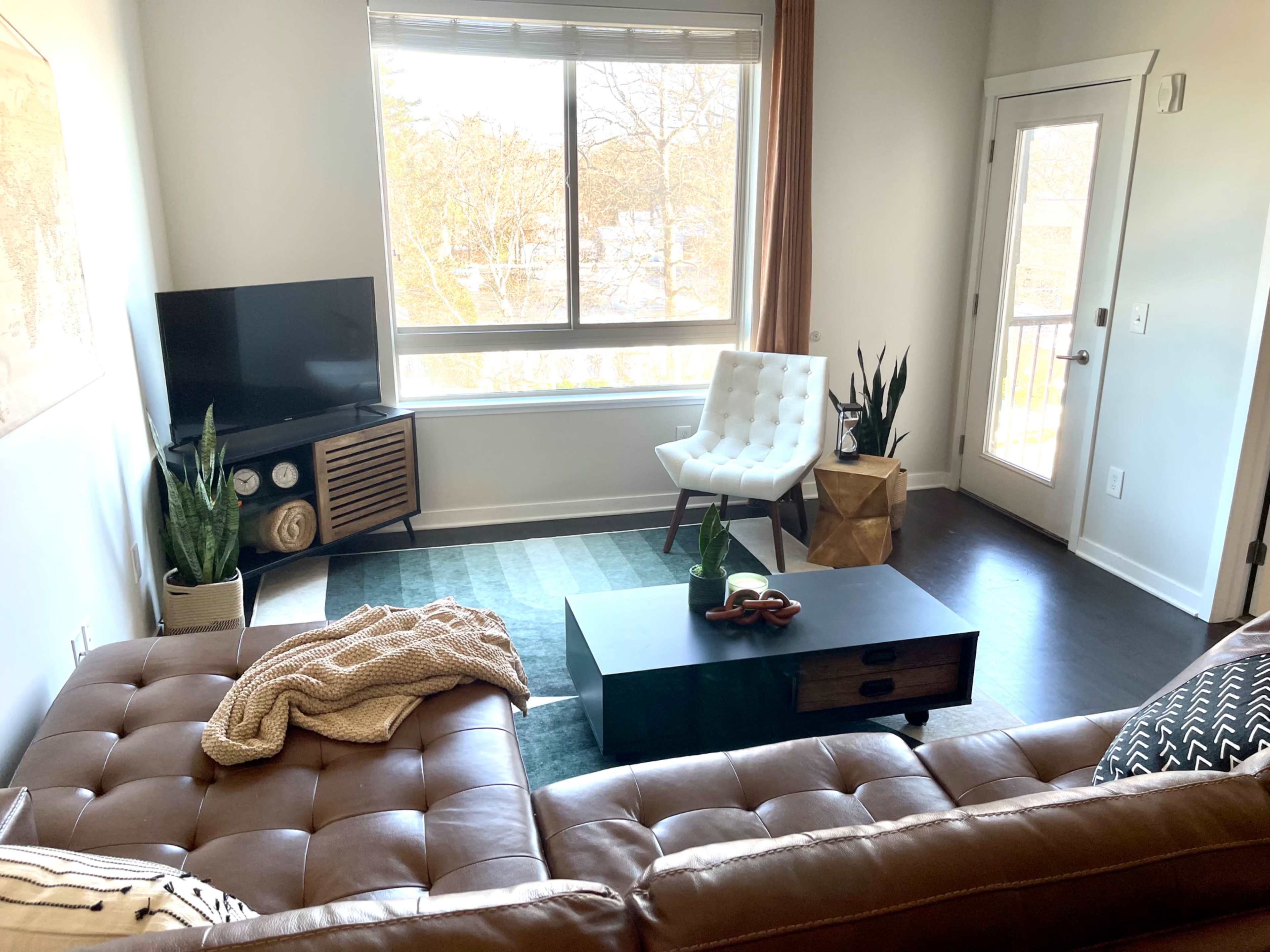 A modern living room with a brown leather sofa, a coffee table, a white chair, a television, and a view of a balcony through large windows.