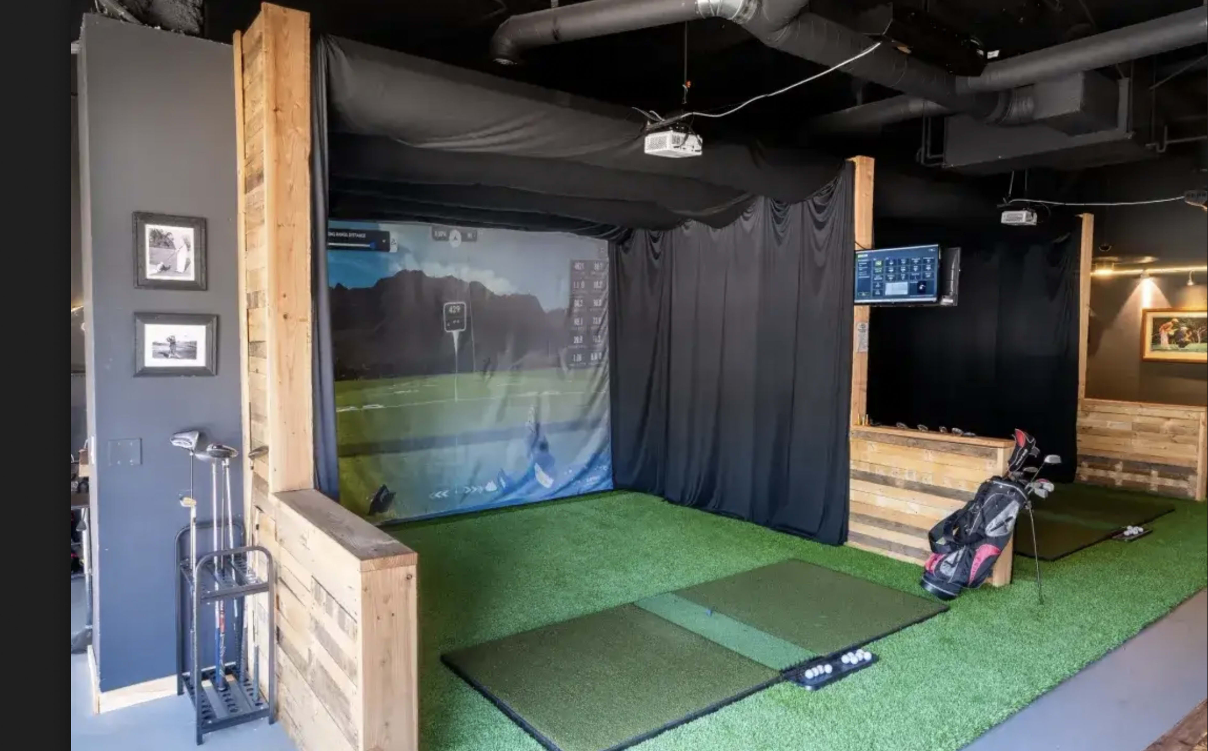 The image shows an indoor golf simulation area featuring a green turf floor, a large projection screen, and golf clubs in a bag situated nearby.