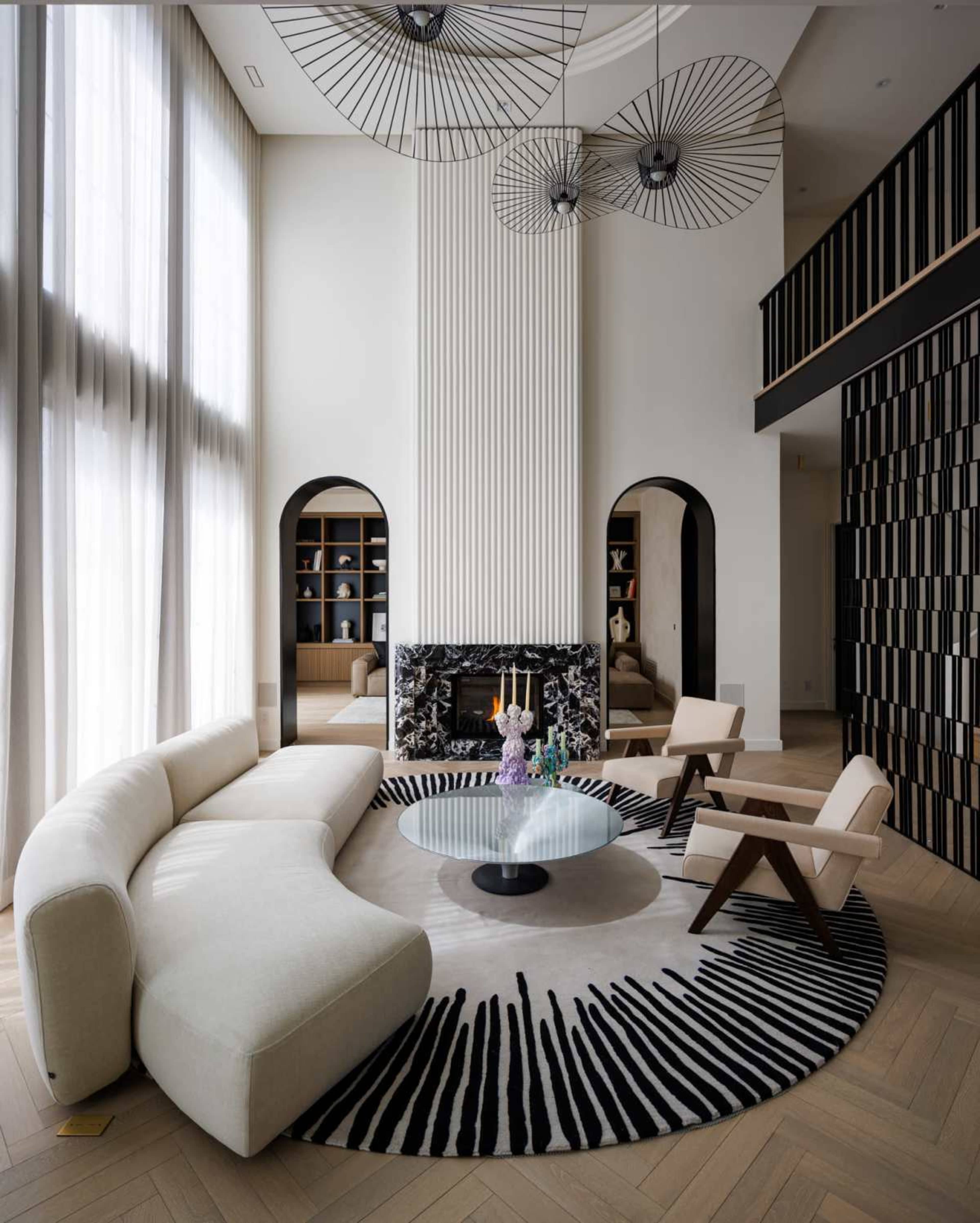 The modern living room features a curved white sofa, a round area rug with a bold pattern, and a marble fireplace, illuminated by large windows and artistic light fixtures.