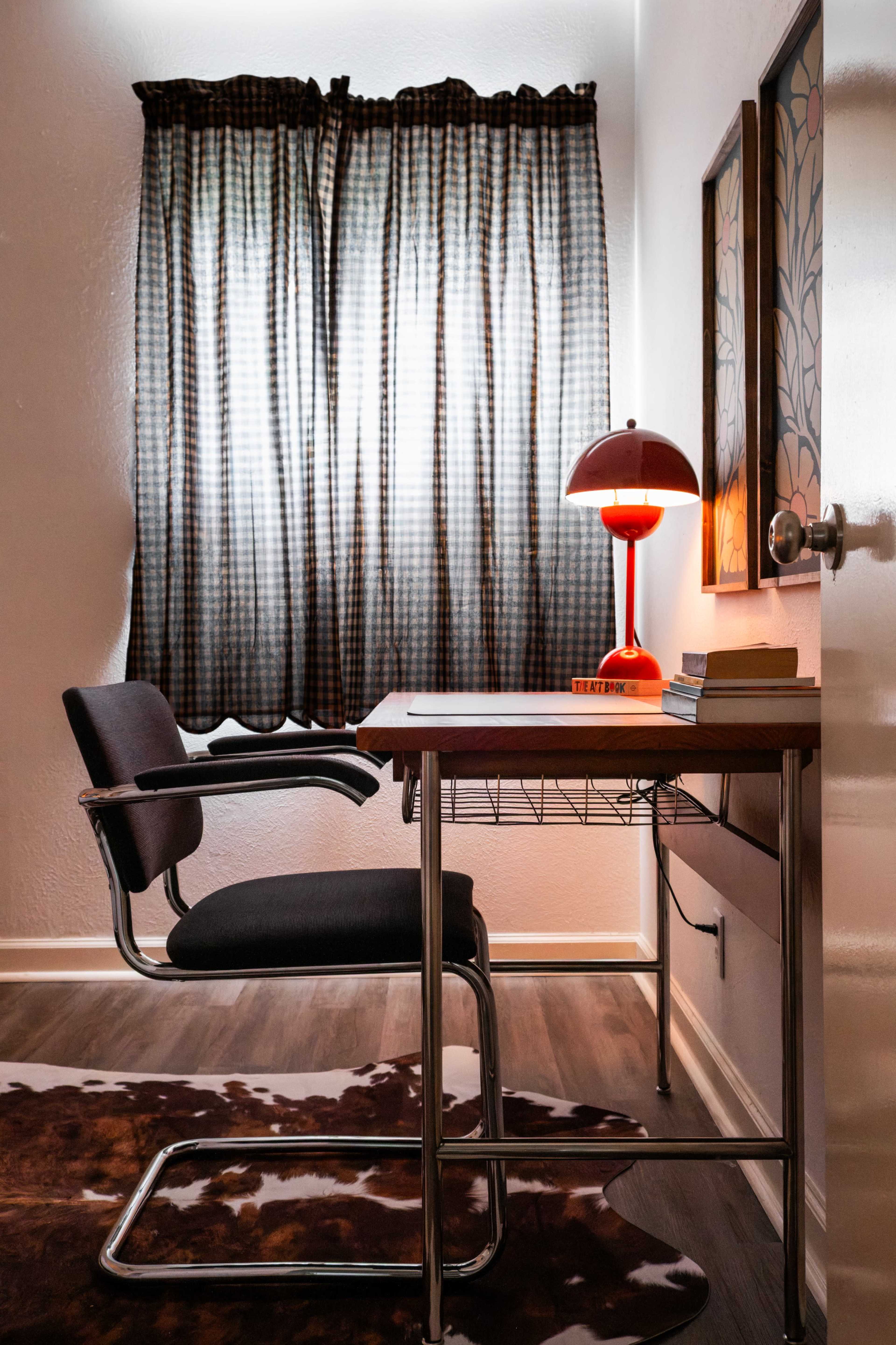A simple desk with a chair is positioned by a window, featuring a lamp and a few books, surrounded by a patterned curtain.