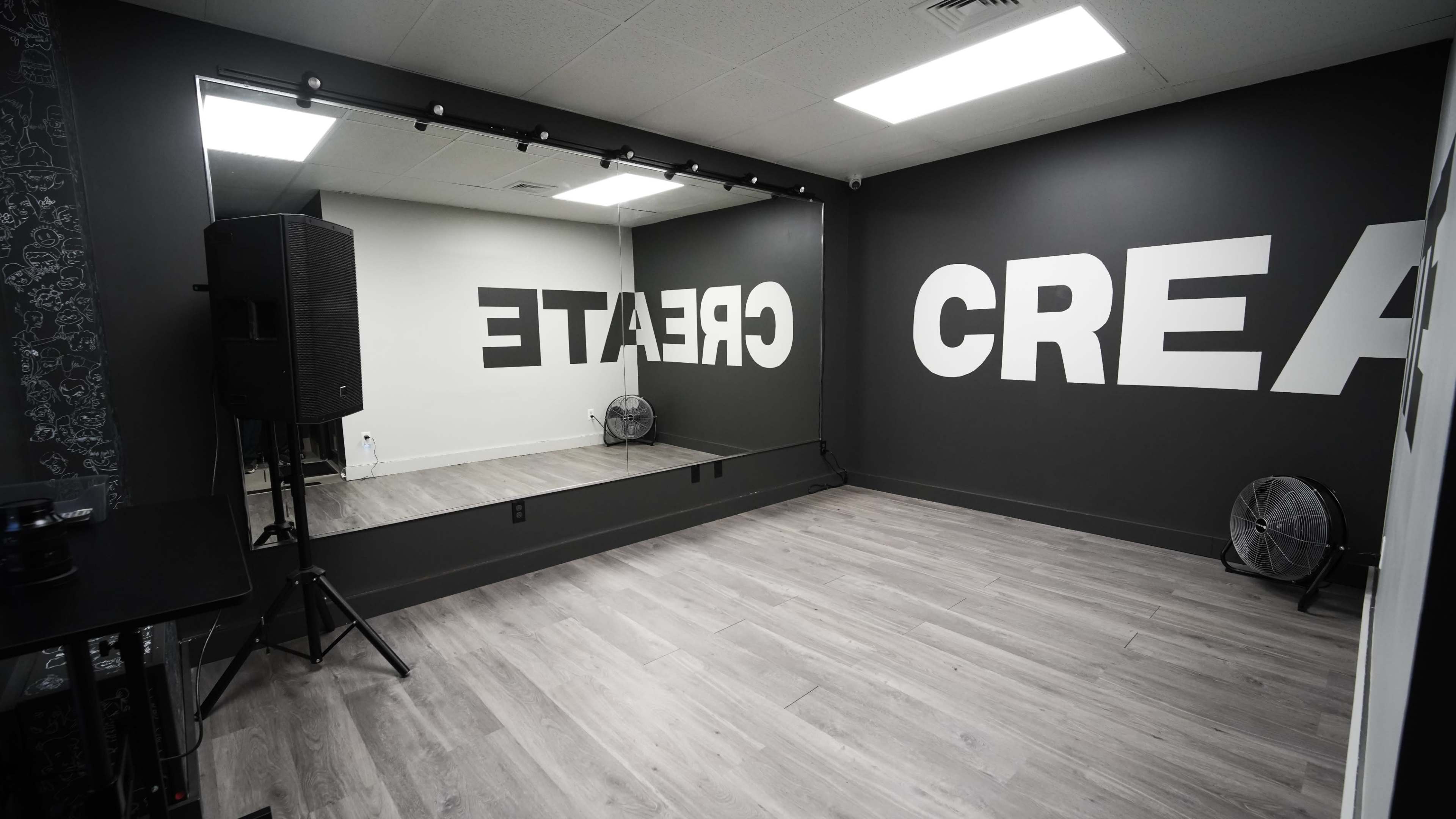 The image shows a spacious room with a large mirror on one wall and the word "CREATE" prominently displayed on another wall, featuring gray walls and wooden flooring.