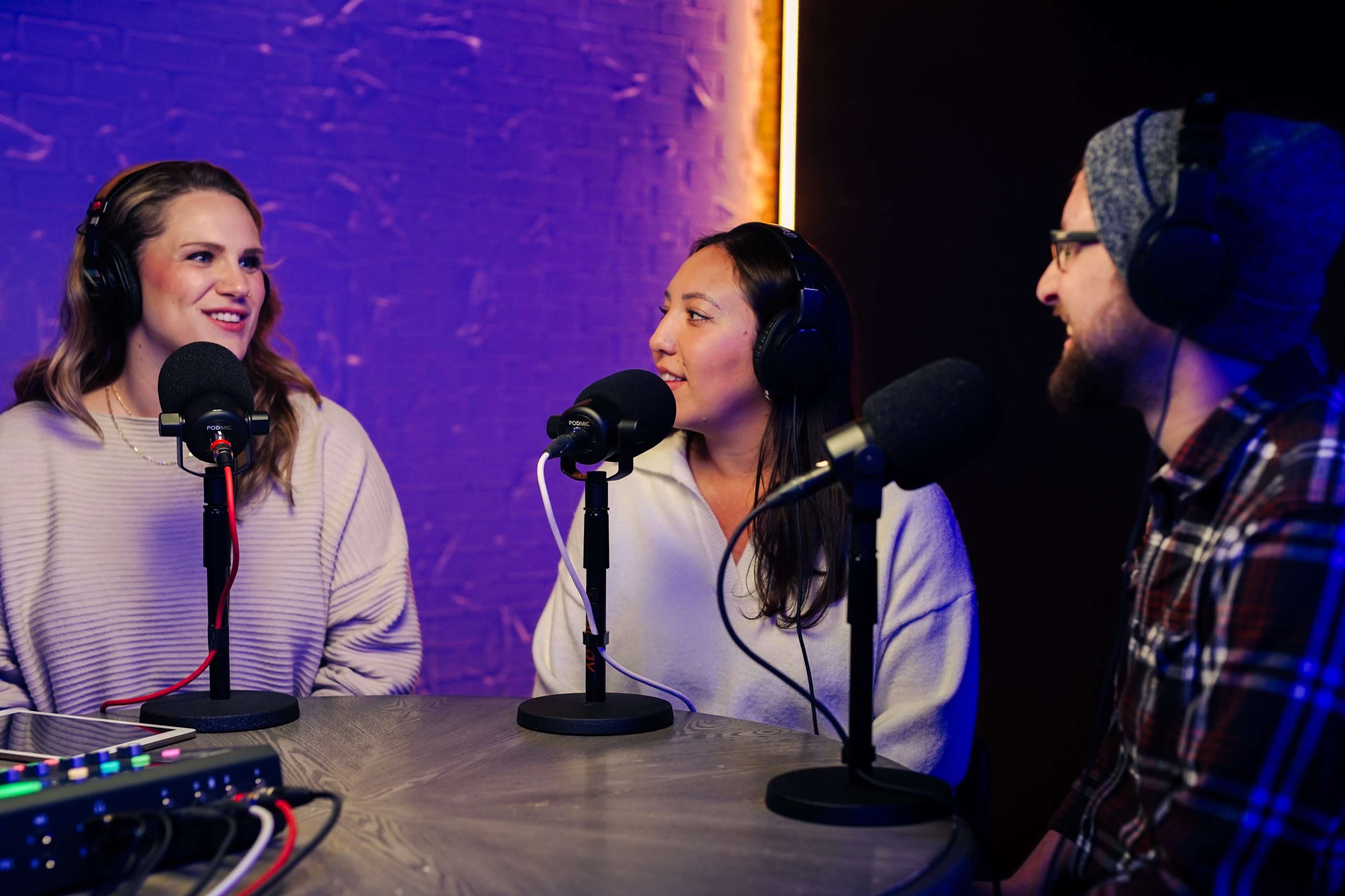 Three individuals are seated around a table with microphones in a brightly lit podcast studio.