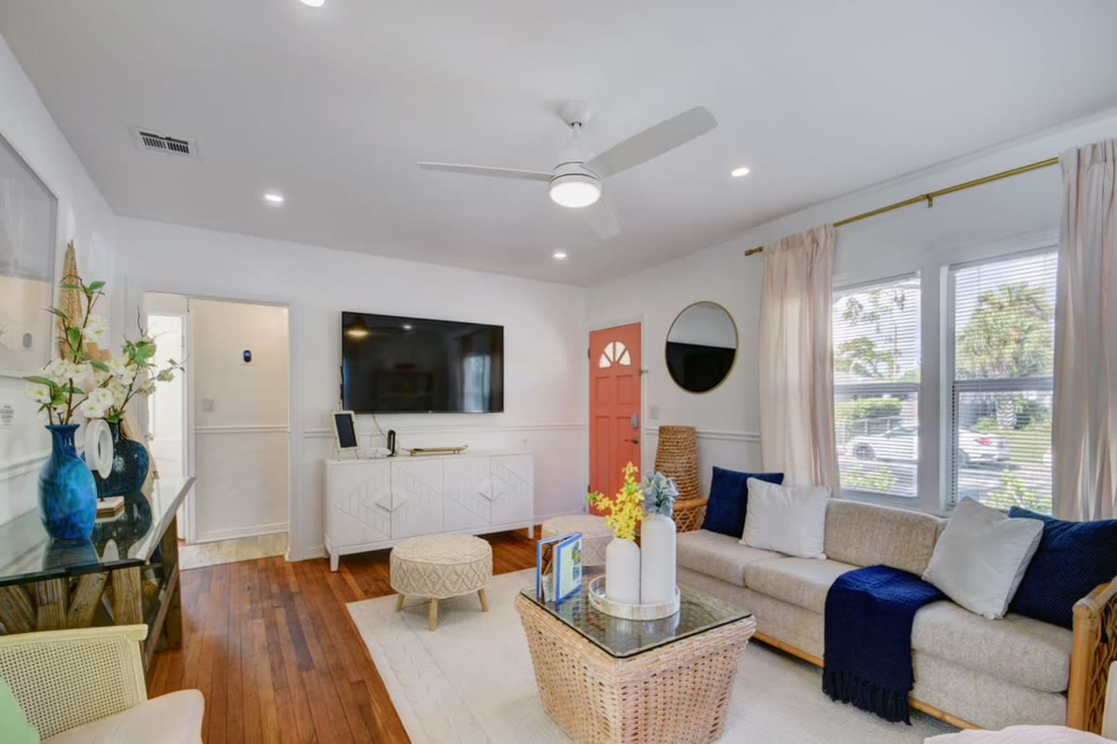 A bright living room features a beige sofa, a large television mounted on the wall, and a pink door with light streaming through the windows.