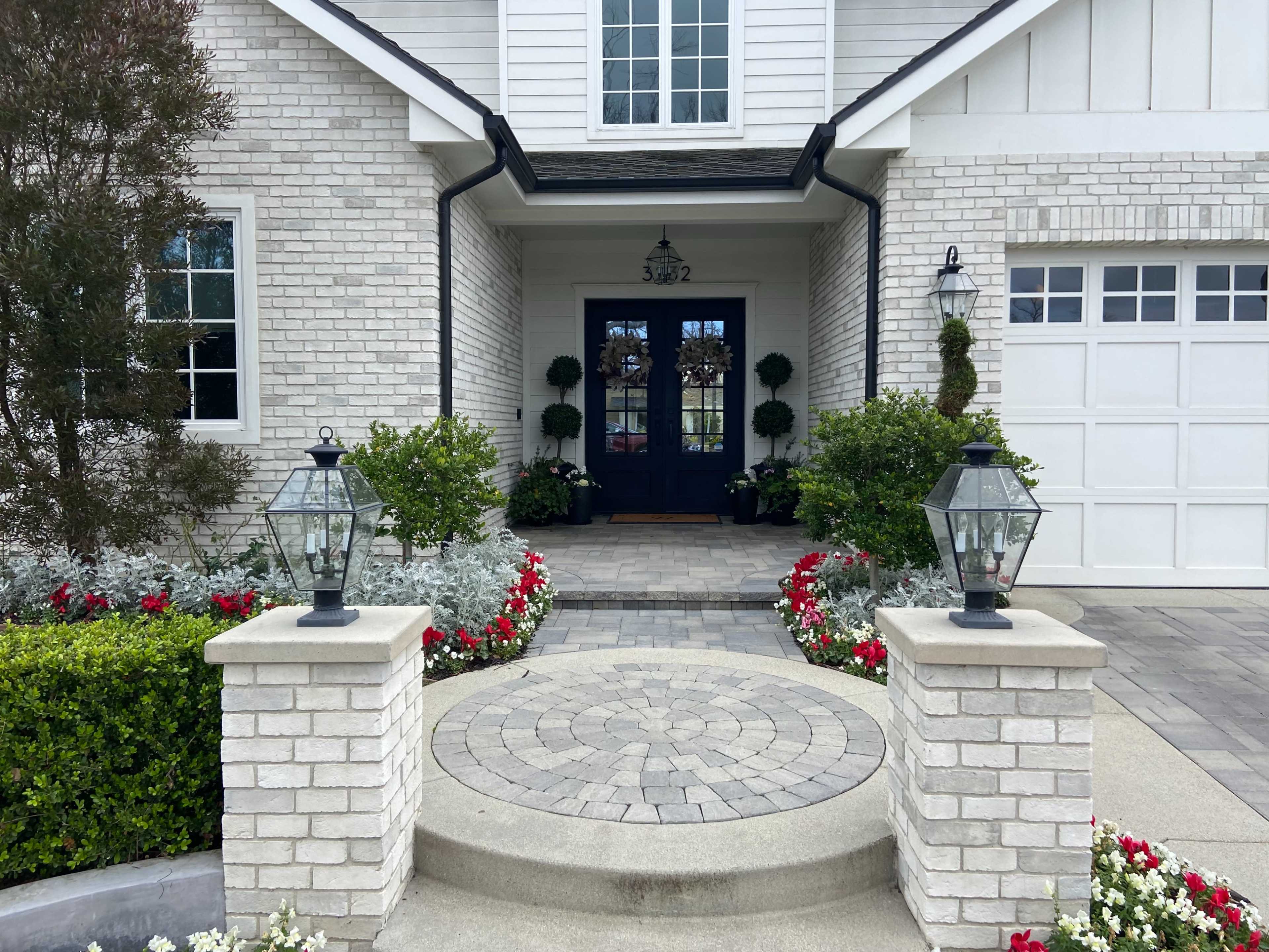 A well-maintained entrance with a circular stone pathway leading to a blue door, flanked by lanterns and surrounded by colorful landscaping.