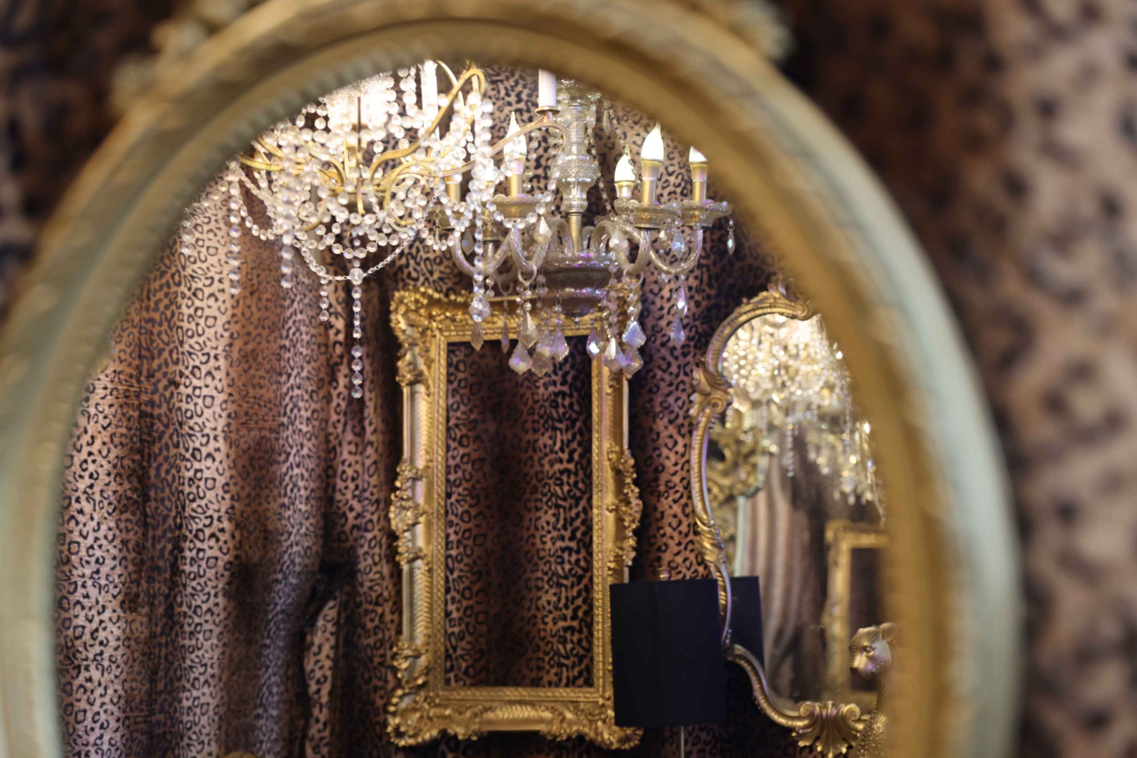The image shows a lavishly decorated room featuring a chandelier, an ornate mirror, and walls covered with a leopard print fabric.