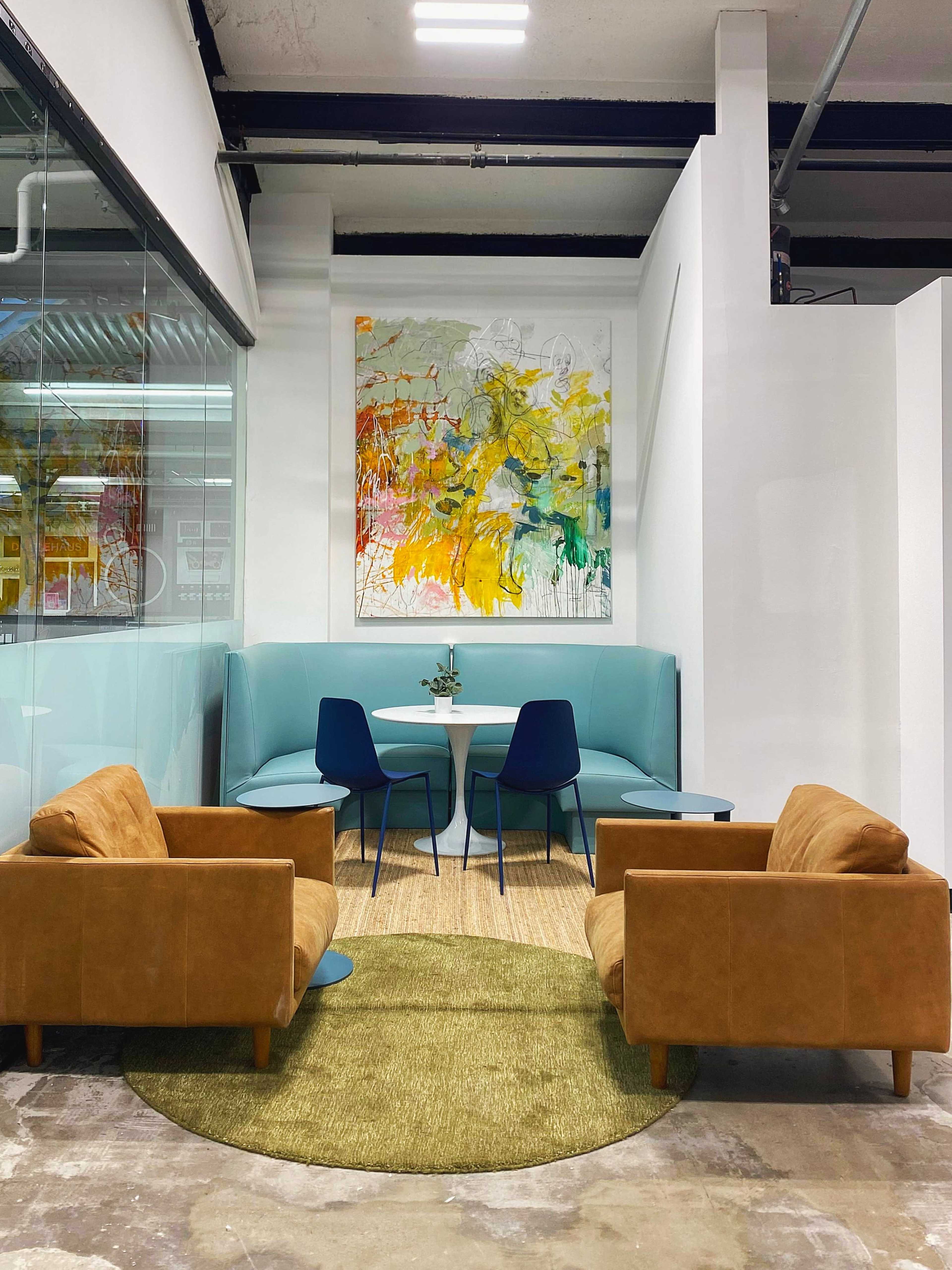 The image shows a modern lounge area featuring two brown armchairs, a light blue booth, a circular table, and a large colorful artwork on the wall.