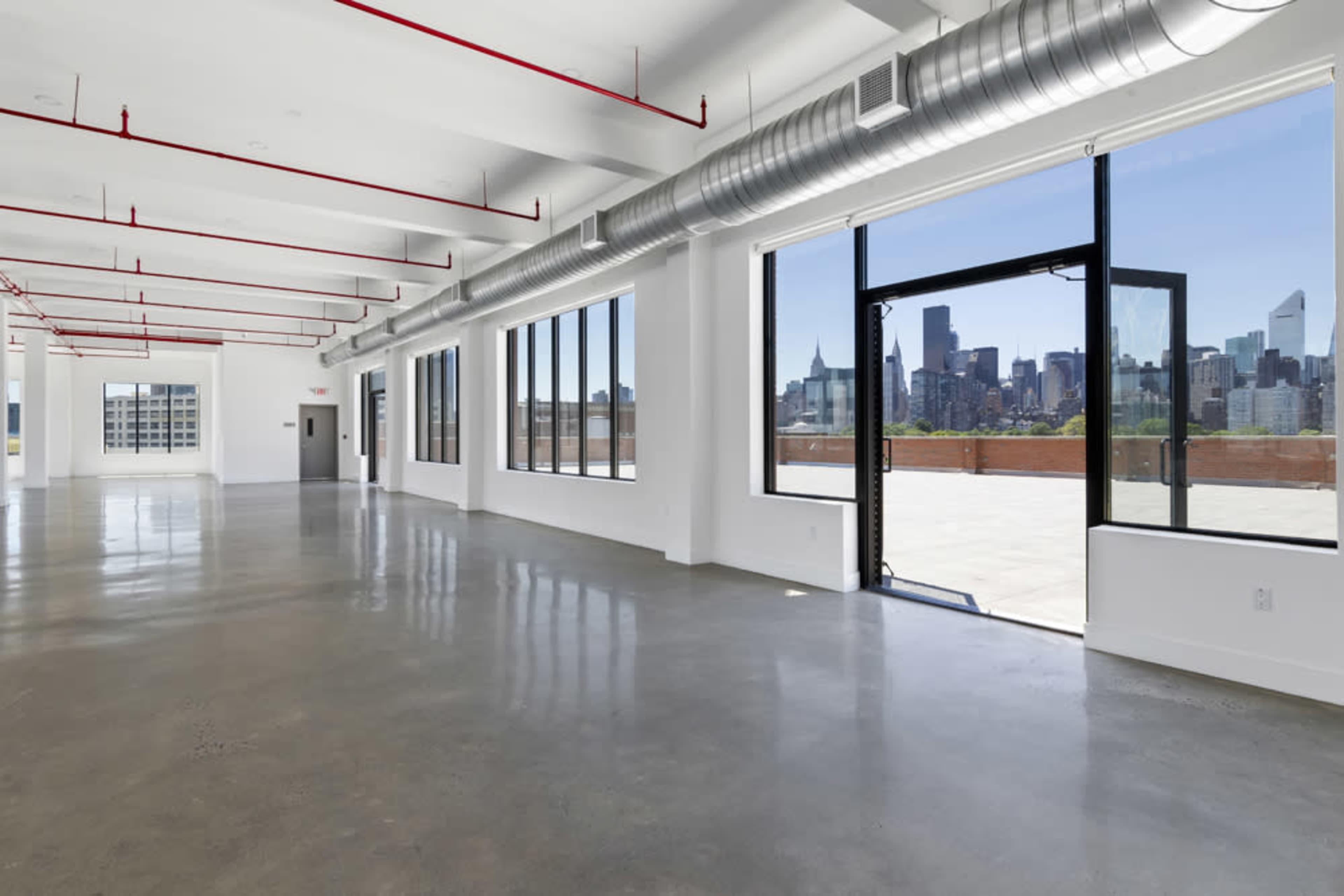 A spacious, empty loft with large windows and a door leading to a terrace, offering a view of a city skyline.