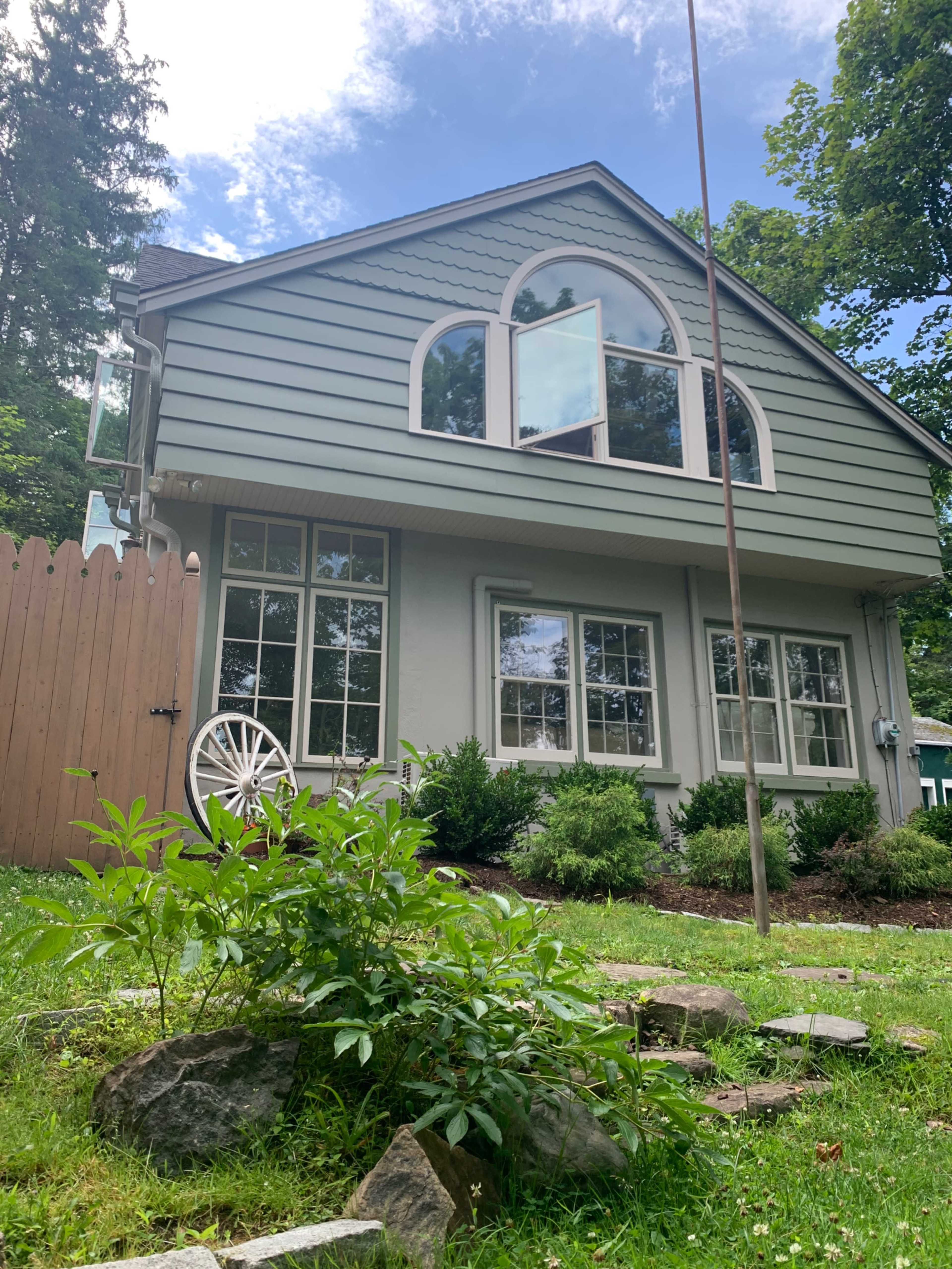 A gray house with large windows and an arched top stands in a grassy area surrounded by trees and stone pathways.