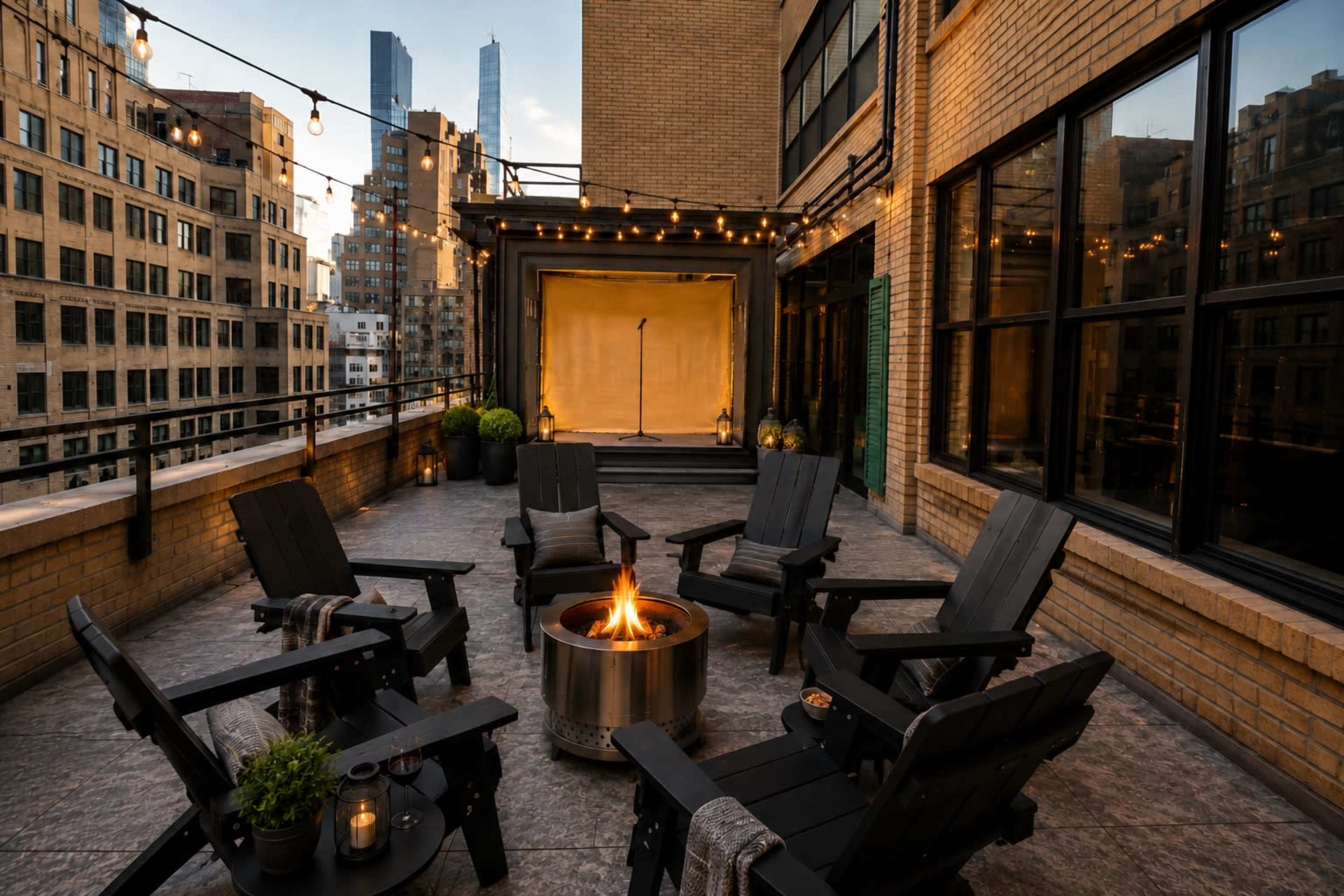 A rooftop terrace features several black chairs arranged around a fire pit, with city buildings and a stage backdrop in the distance.